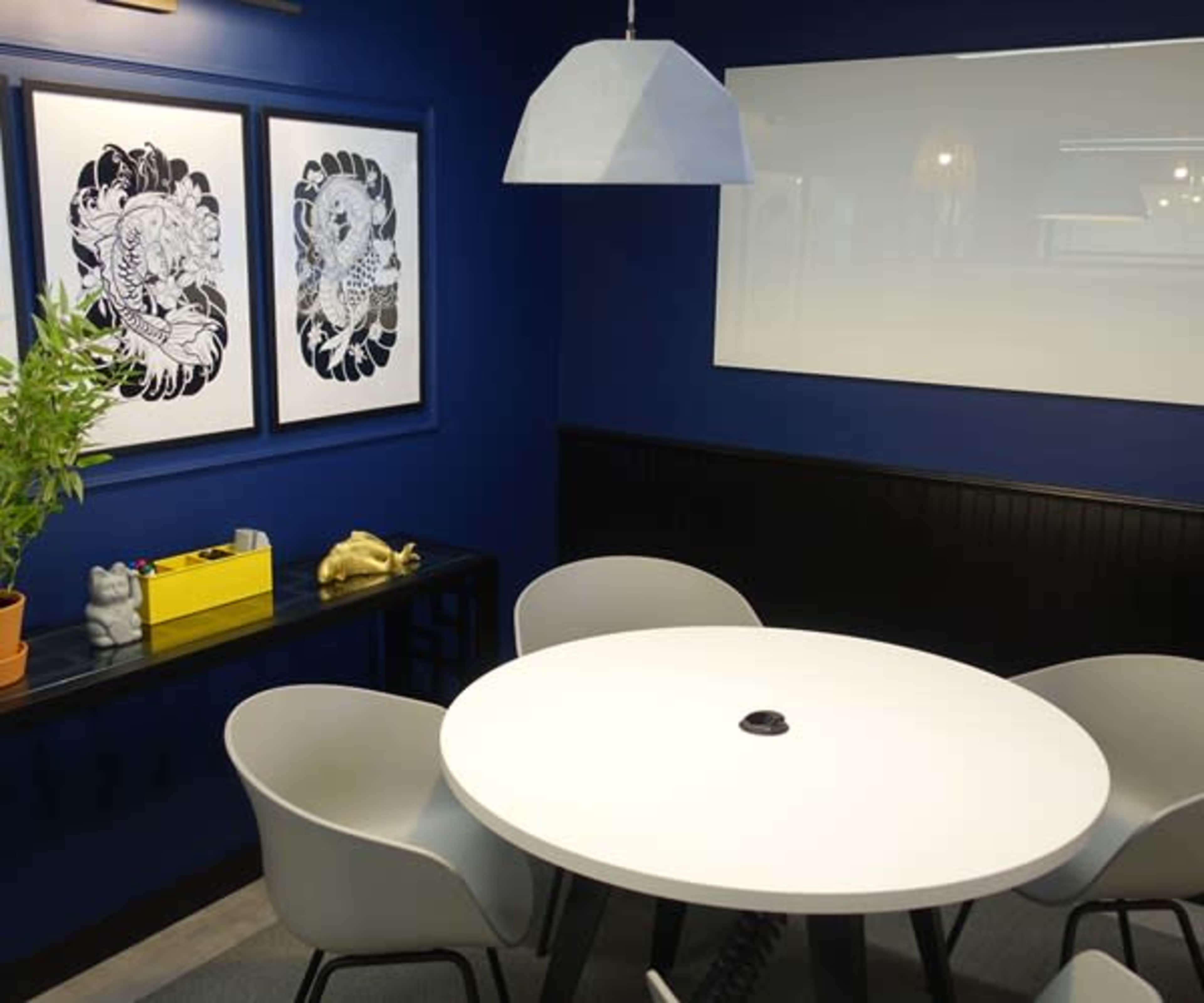A modern meeting room featuring a round white table surrounded by four gray chairs, with dark blue walls adorned by two black-and-white framed artworks, and a hanging lamp above the table.