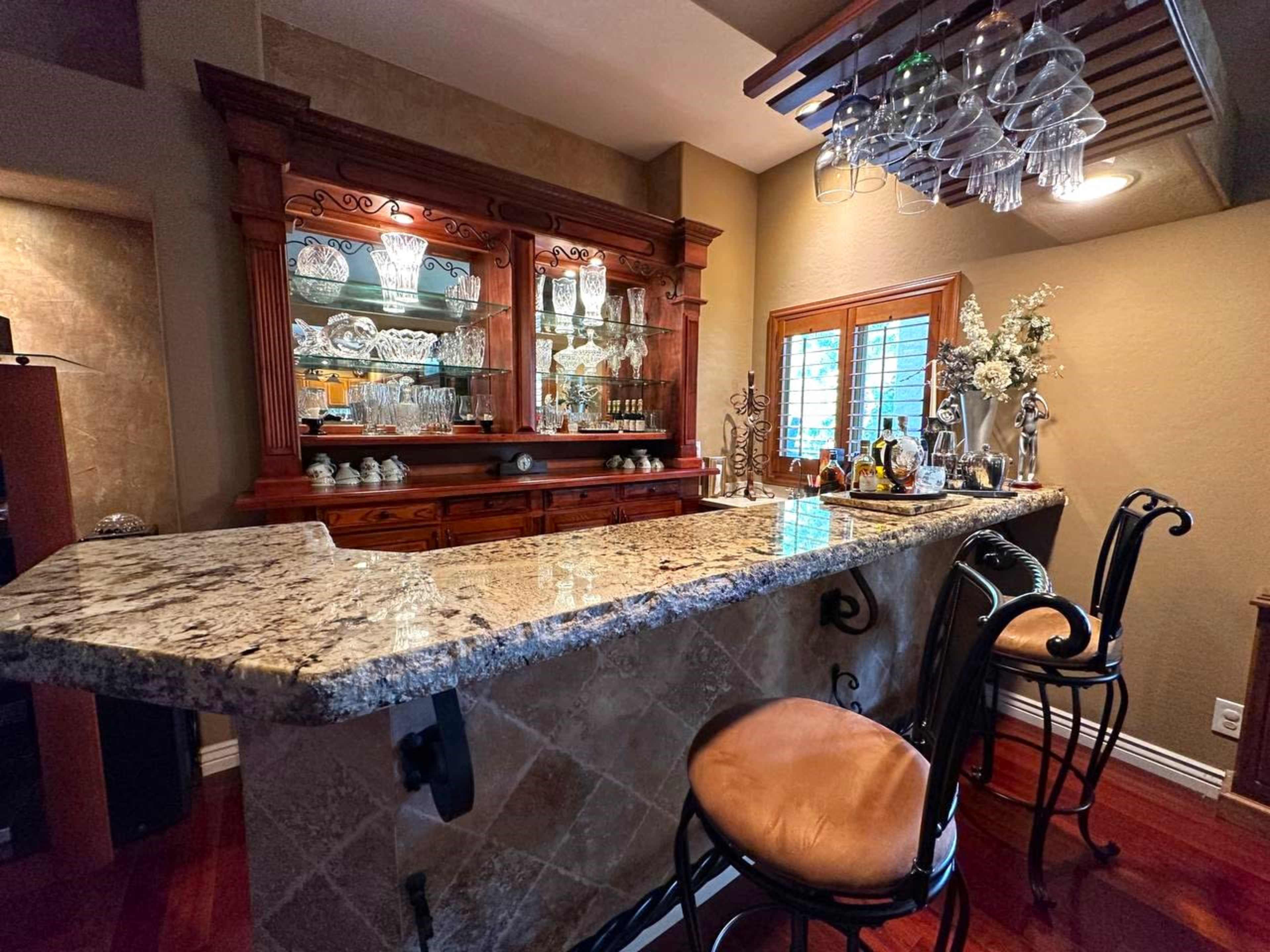 A home bar area with a granite countertop, wooden cabinetry, and a display of glassware under a hanging light fixture.