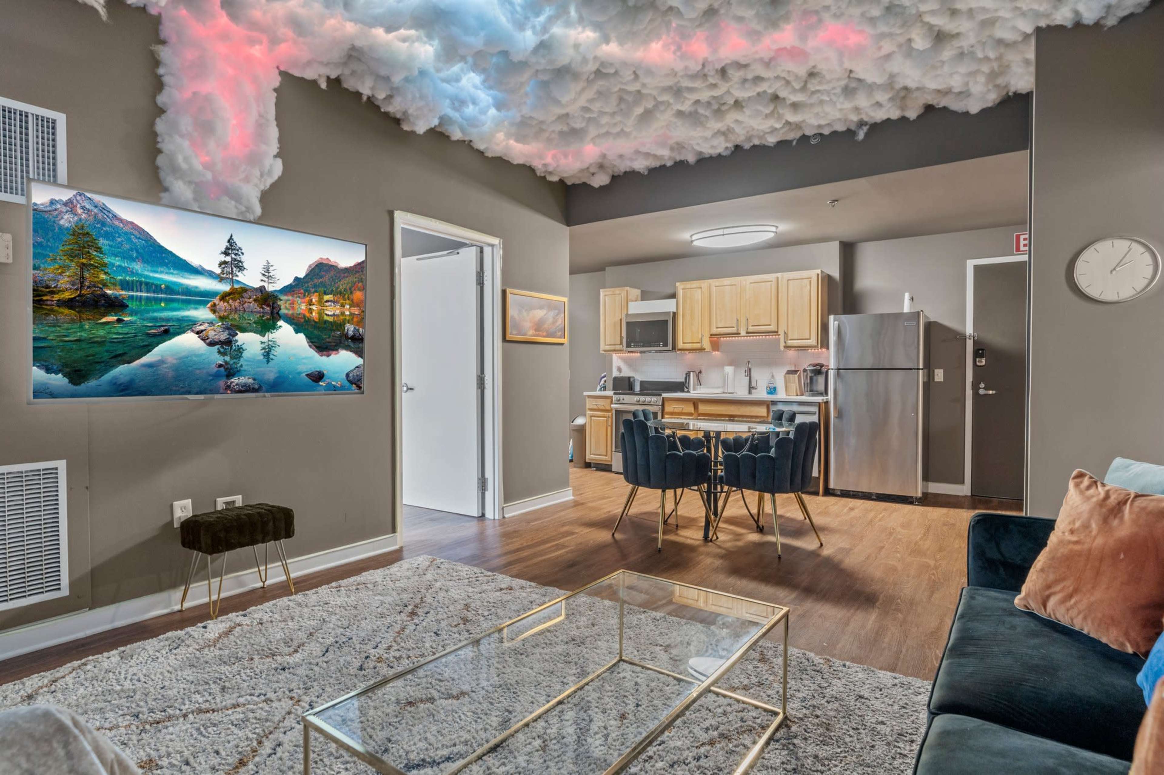 A modern living space with a kitchen area, a television mounted on the wall, and a ceiling designed to resemble fluffy clouds.