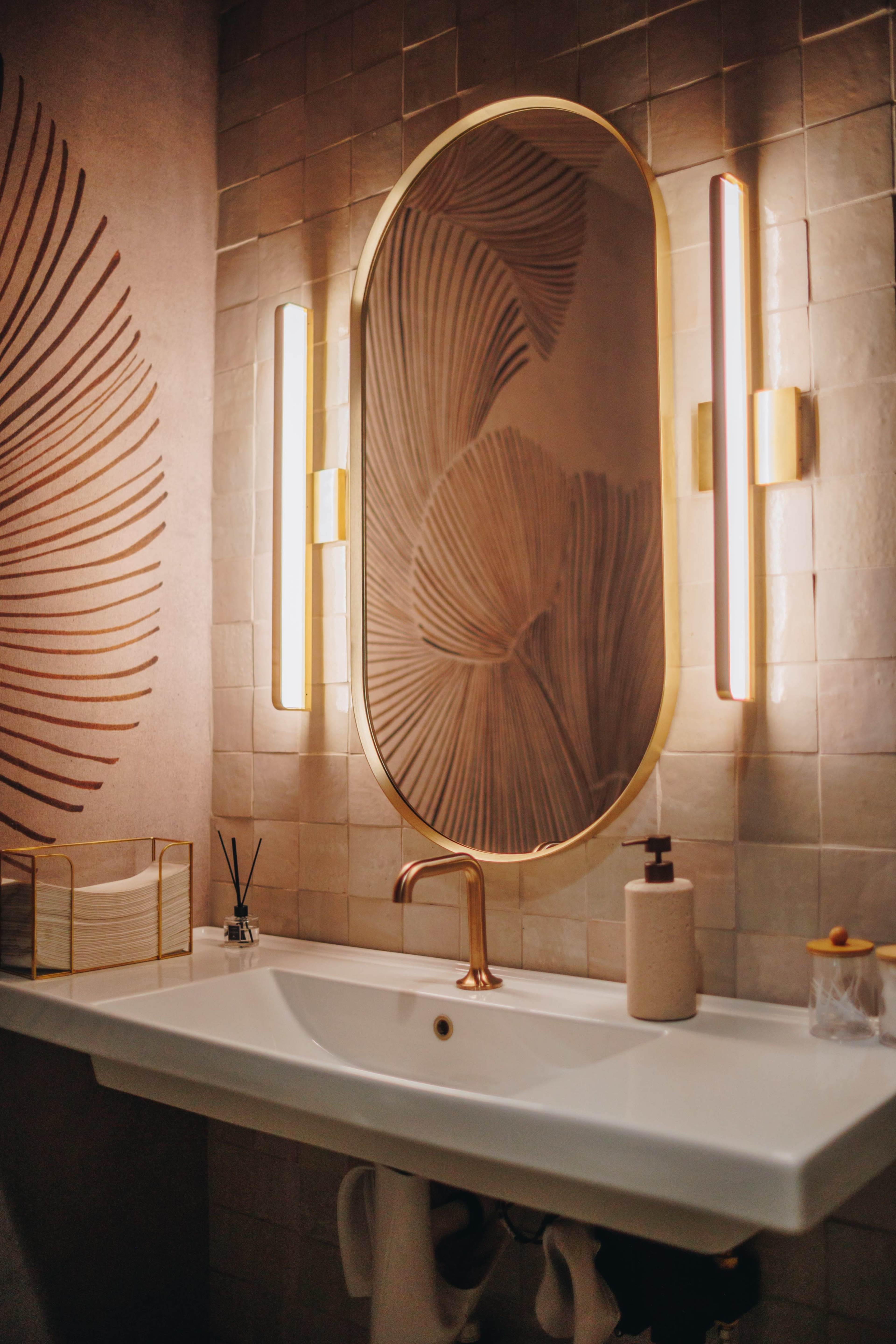 The image shows a modern bathroom with an oval mirror, elegant lighting fixtures, and a sleek sink against a textured wall featuring leaf patterns.