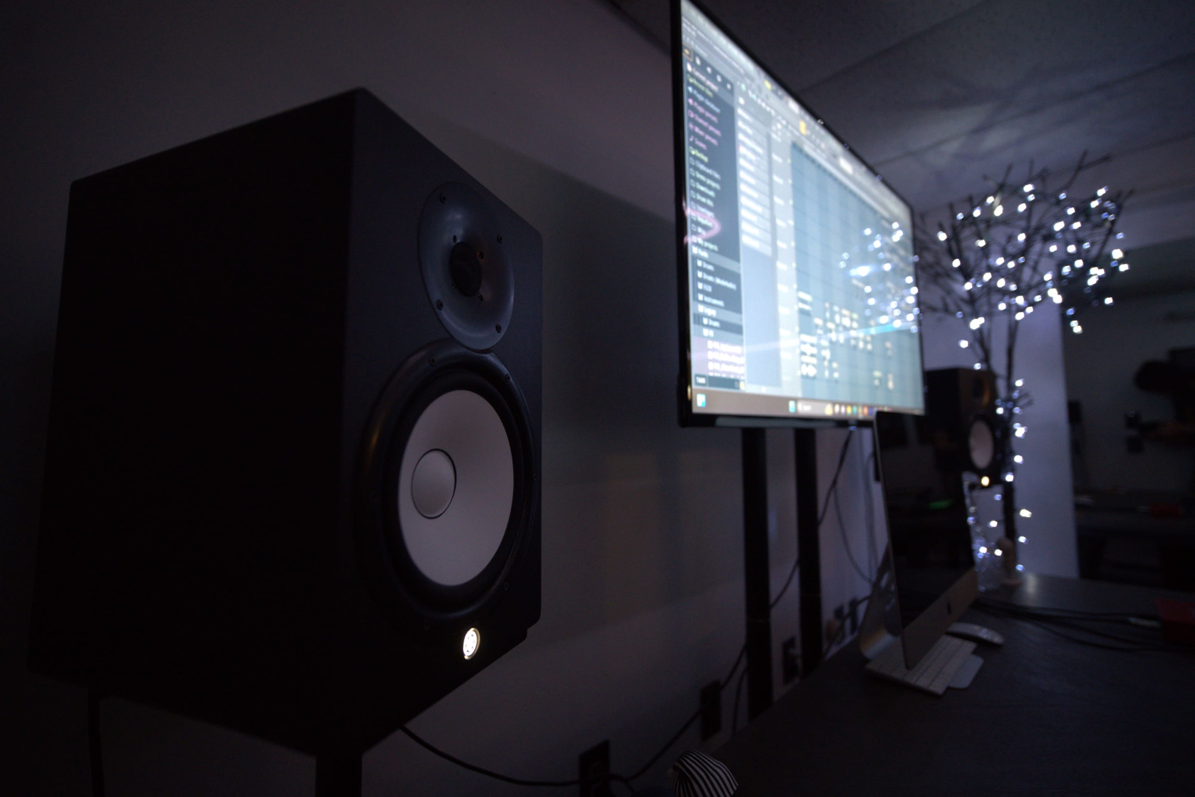A studio monitor speaker stands next to a large screen displaying music software, illuminated by soft lights in the background.