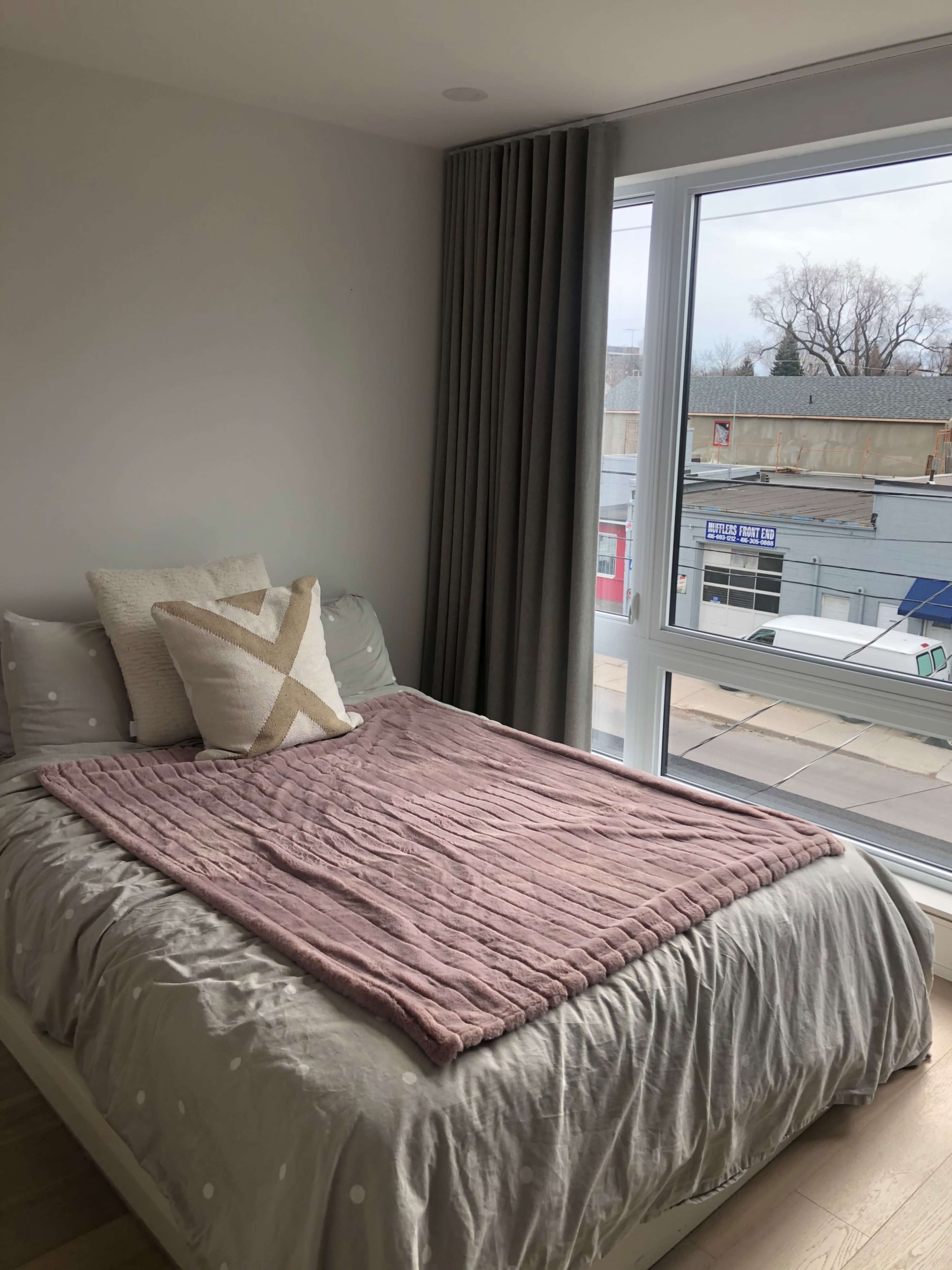 A bed with a pink throw blanket and decorative pillows is positioned next to a large window that overlooks a street.