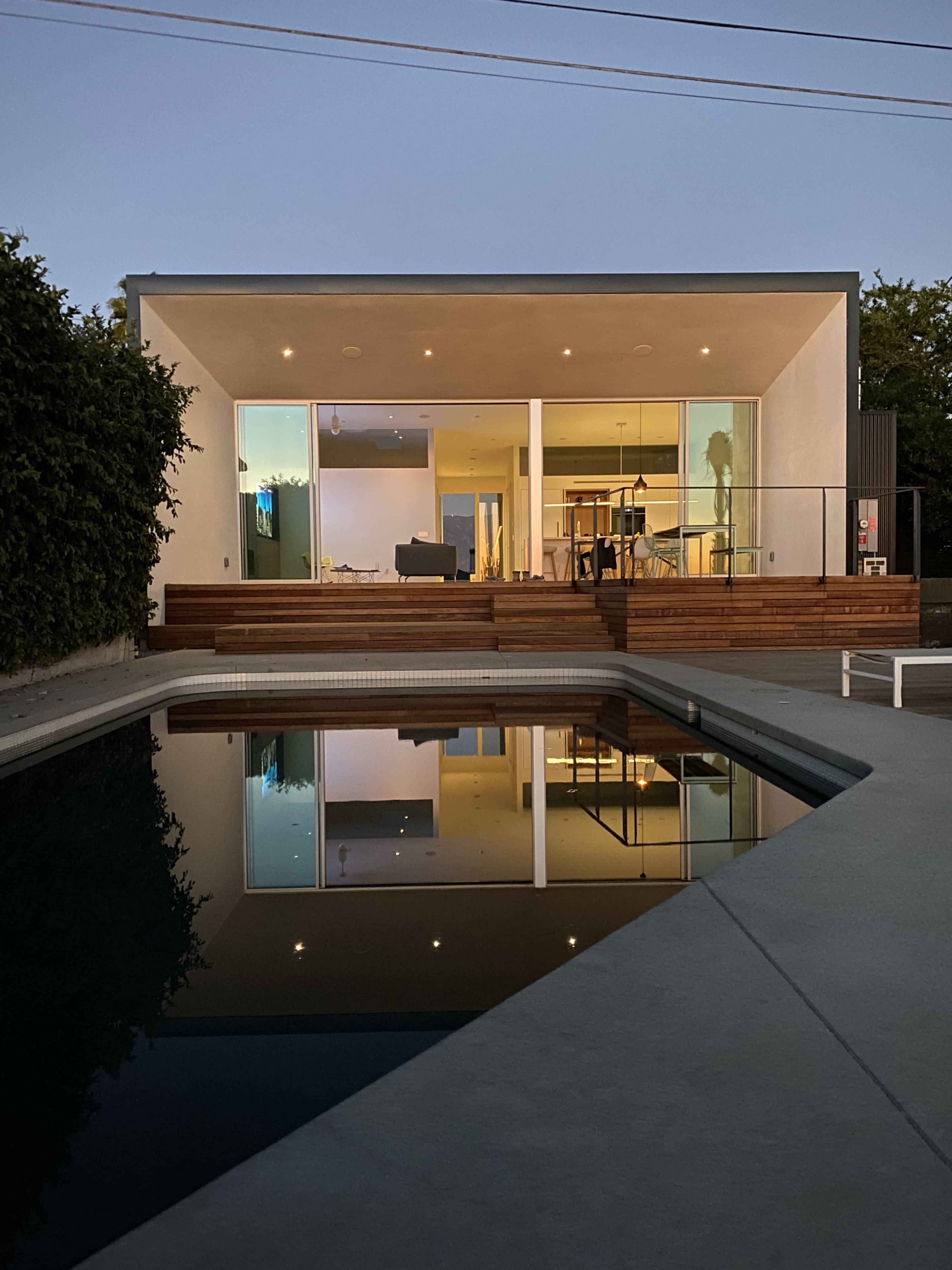 A modern house with large glass windows overlooks a swimming pool, reflecting its exterior and the evening sky.