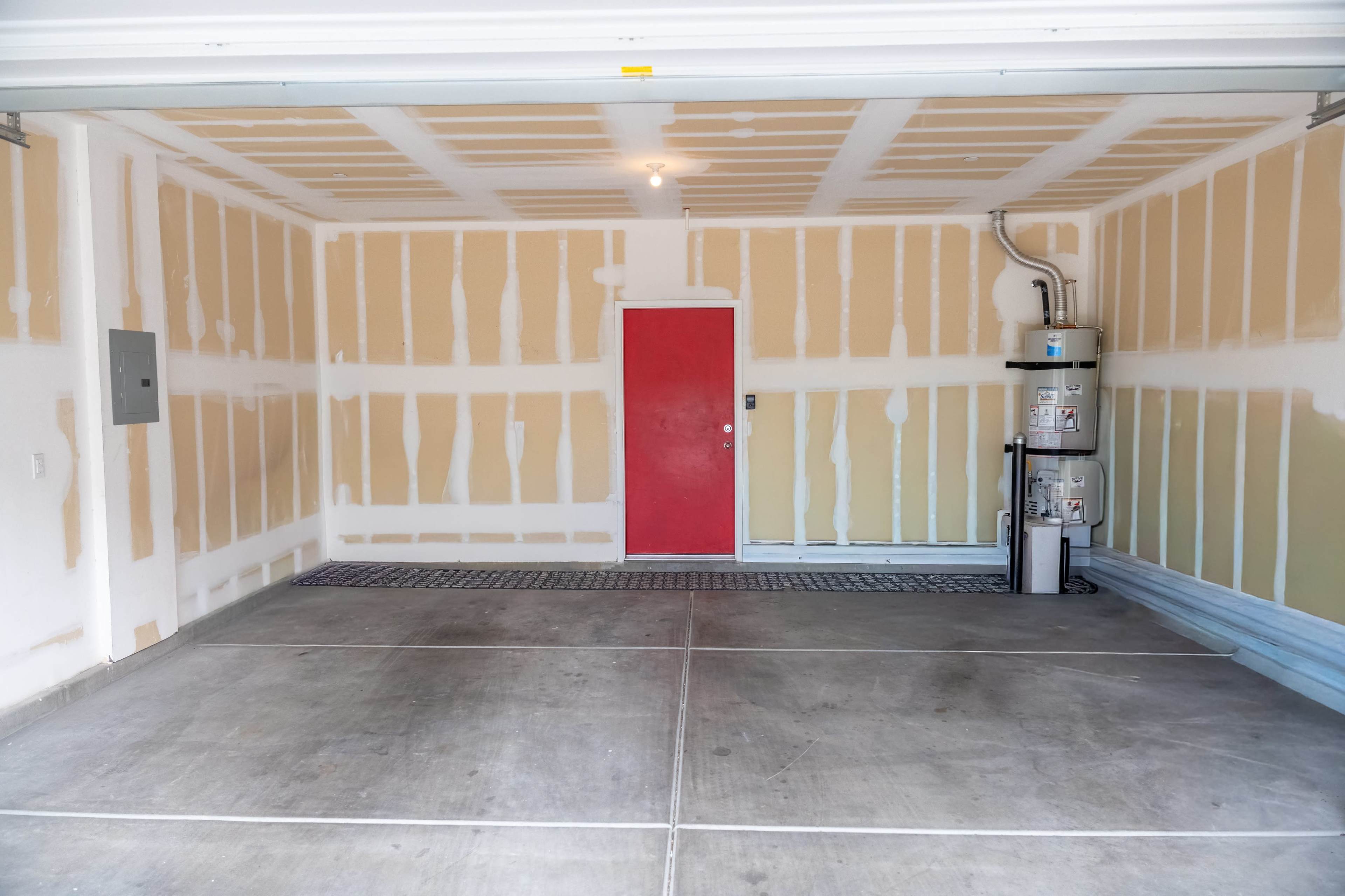The image shows an empty garage with unfinished walls, a red door, and a water heater on the side.