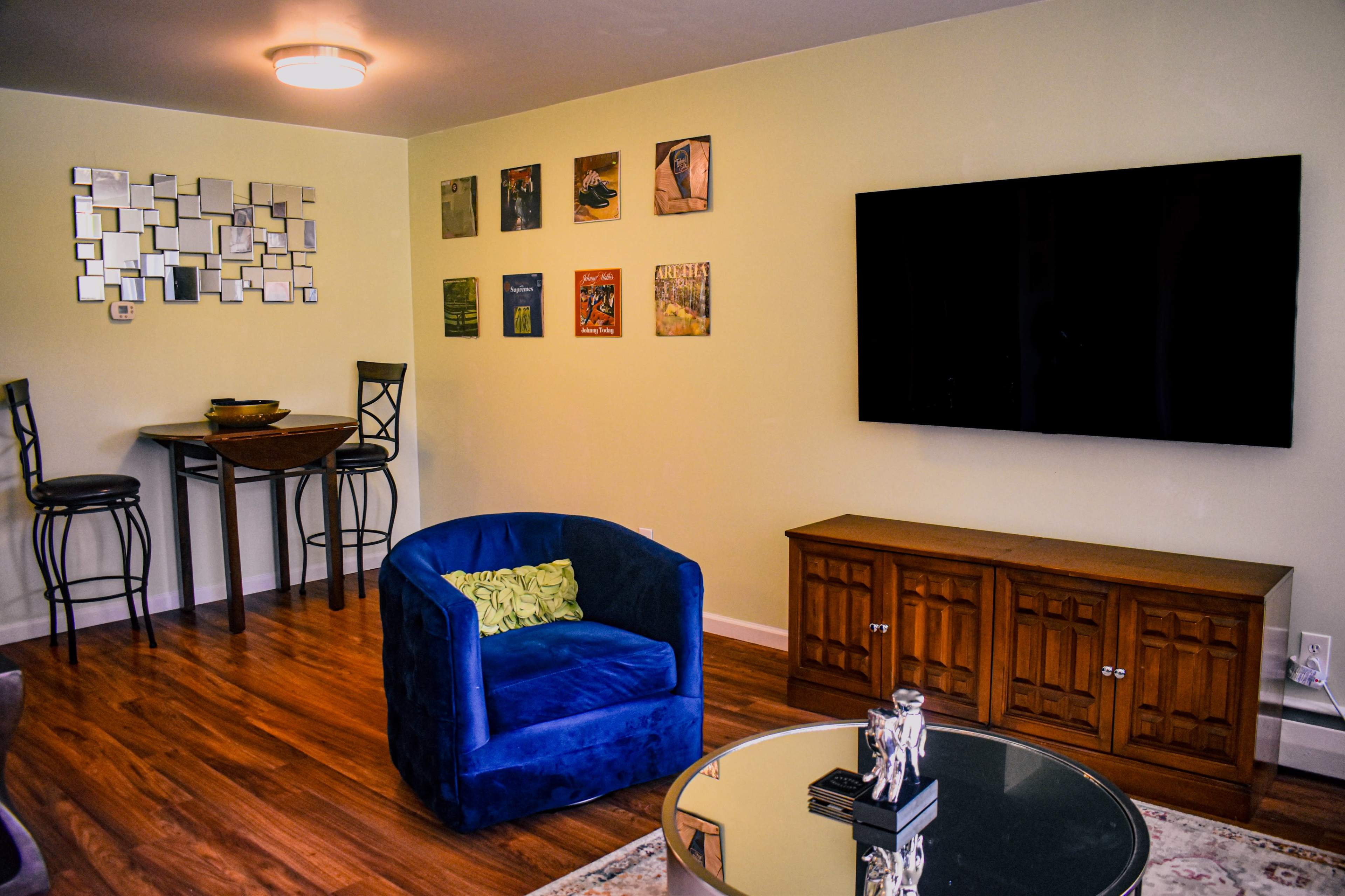 The image shows a living room with a blue armchair, a wooden cabinet, a black flat-screen TV, and a wall decorated with framed artwork and a mirror.