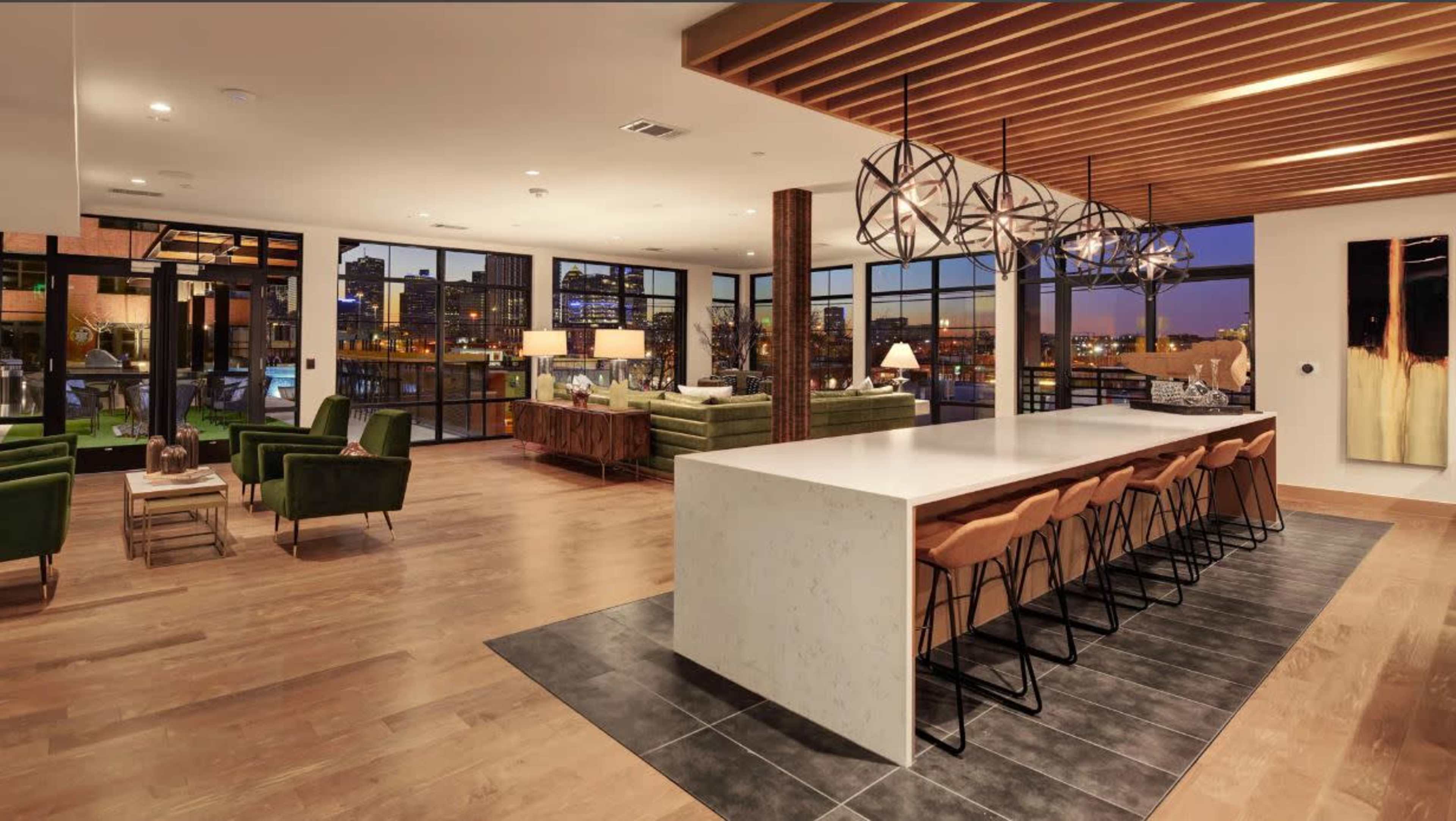A modern interior space featuring a large white kitchen island with bar seating, green chairs, and large windows revealing a city view at dusk.