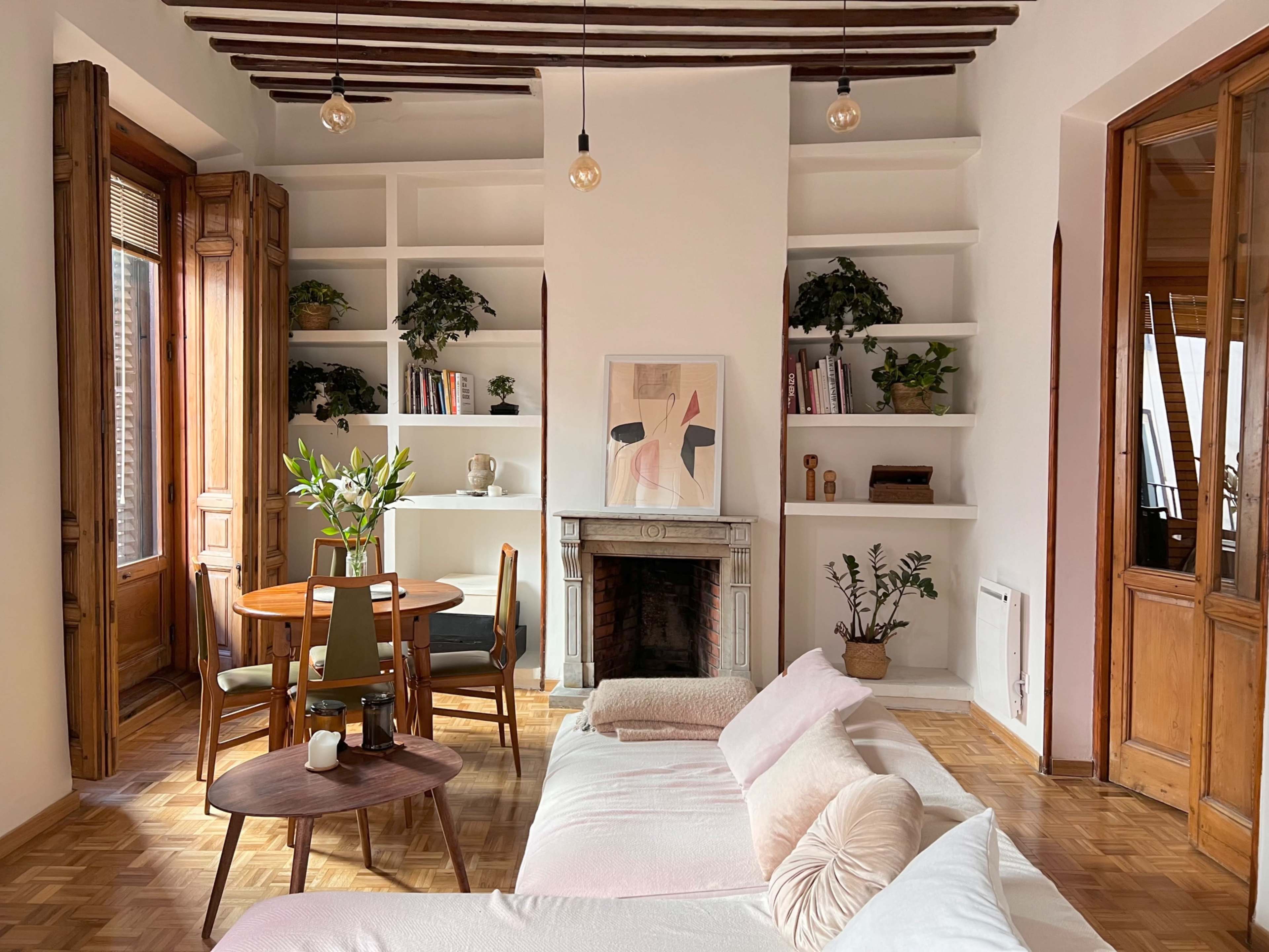 A cozy living room features a white sofa, a wooden dining table with chairs, built-in shelves filled with plants and books, and large windows allowing natural light.