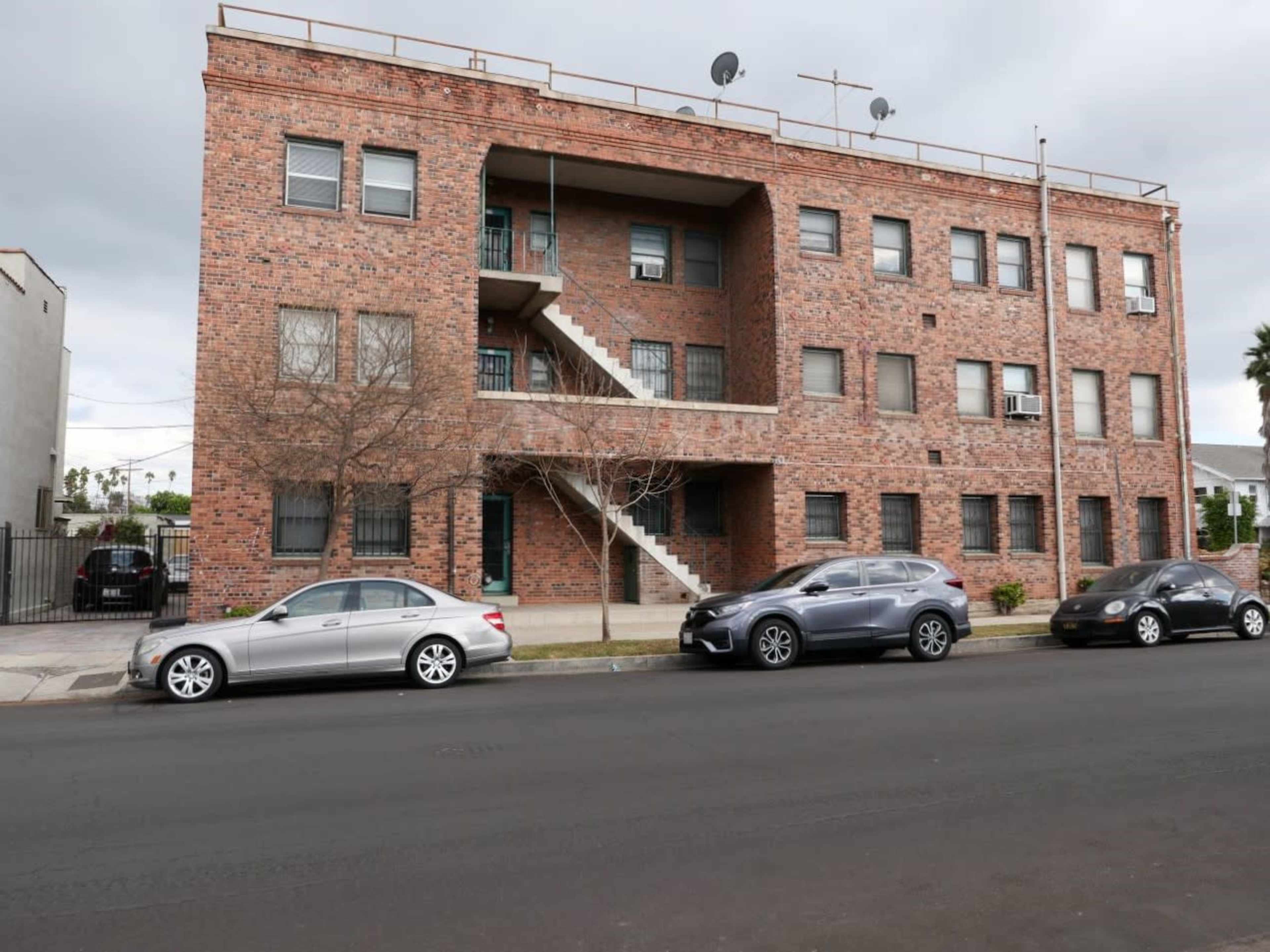 The image shows a three-story brick apartment building with a central staircase and several parked cars along the street in front.