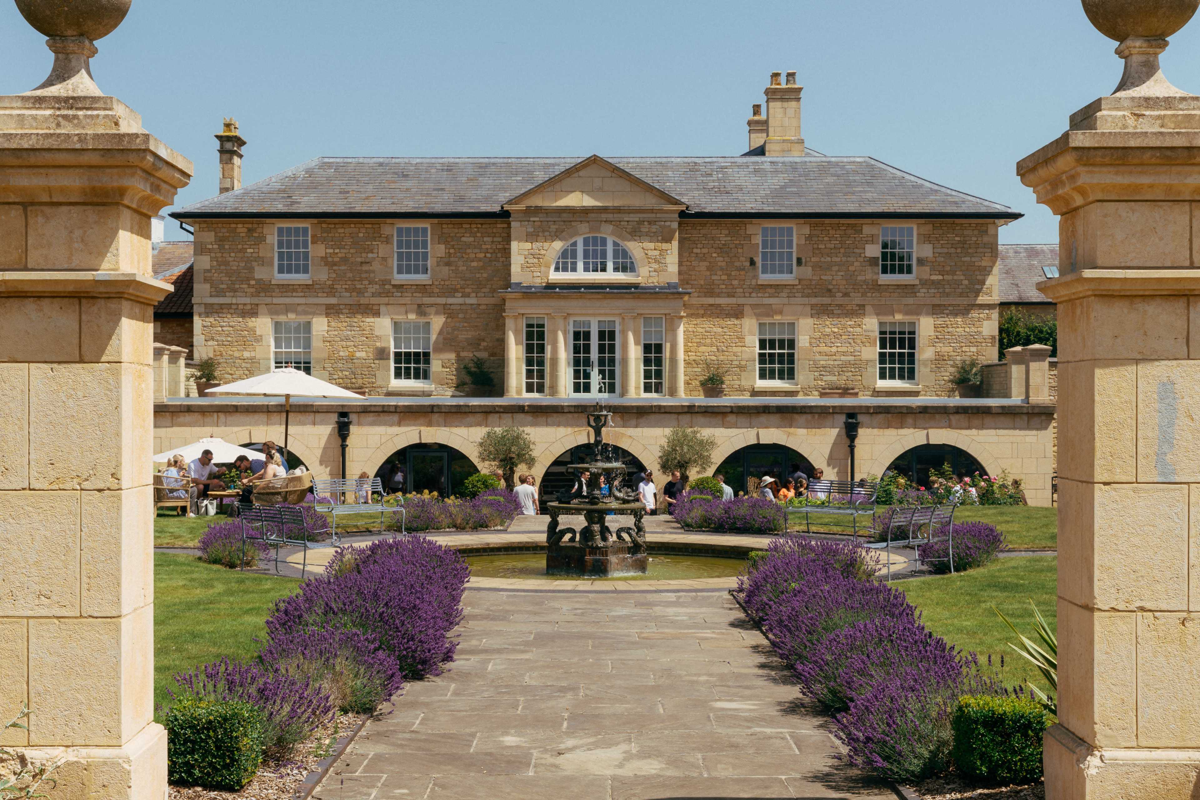 A large stone mansion with arched windows and columns is set behind a fountain surrounded by a garden of blooming lavender.