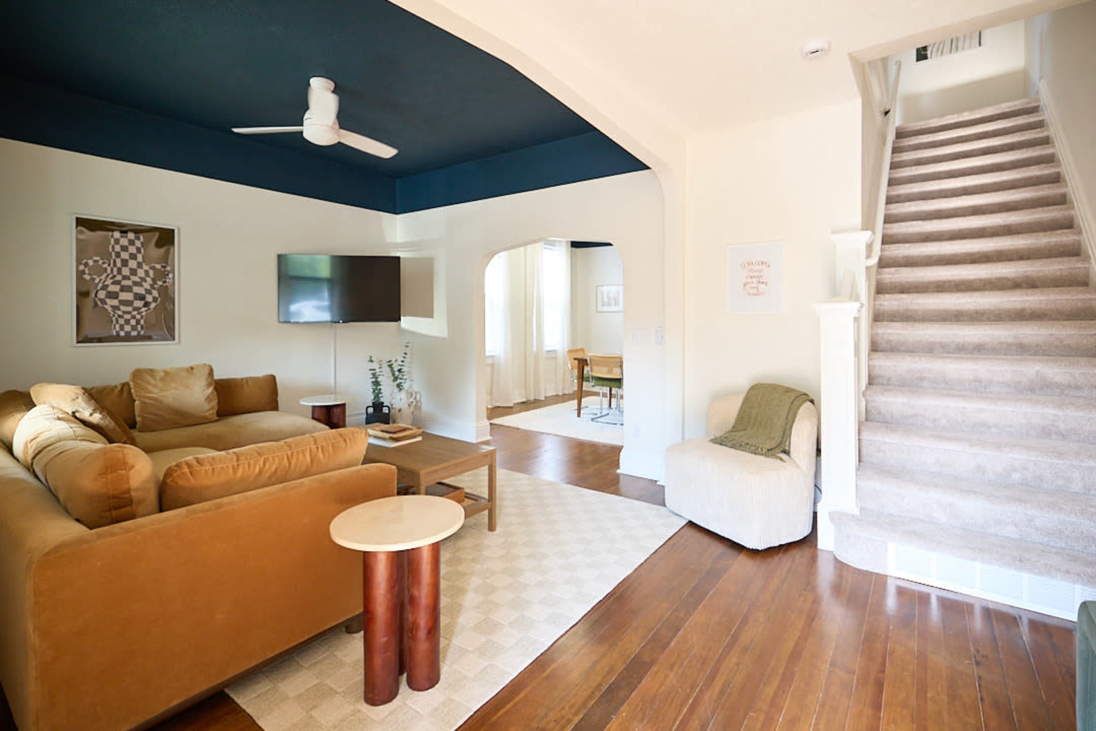 The image shows a living room with a brown sectional sofa, a small round table, and a staircase leading to an upper level.