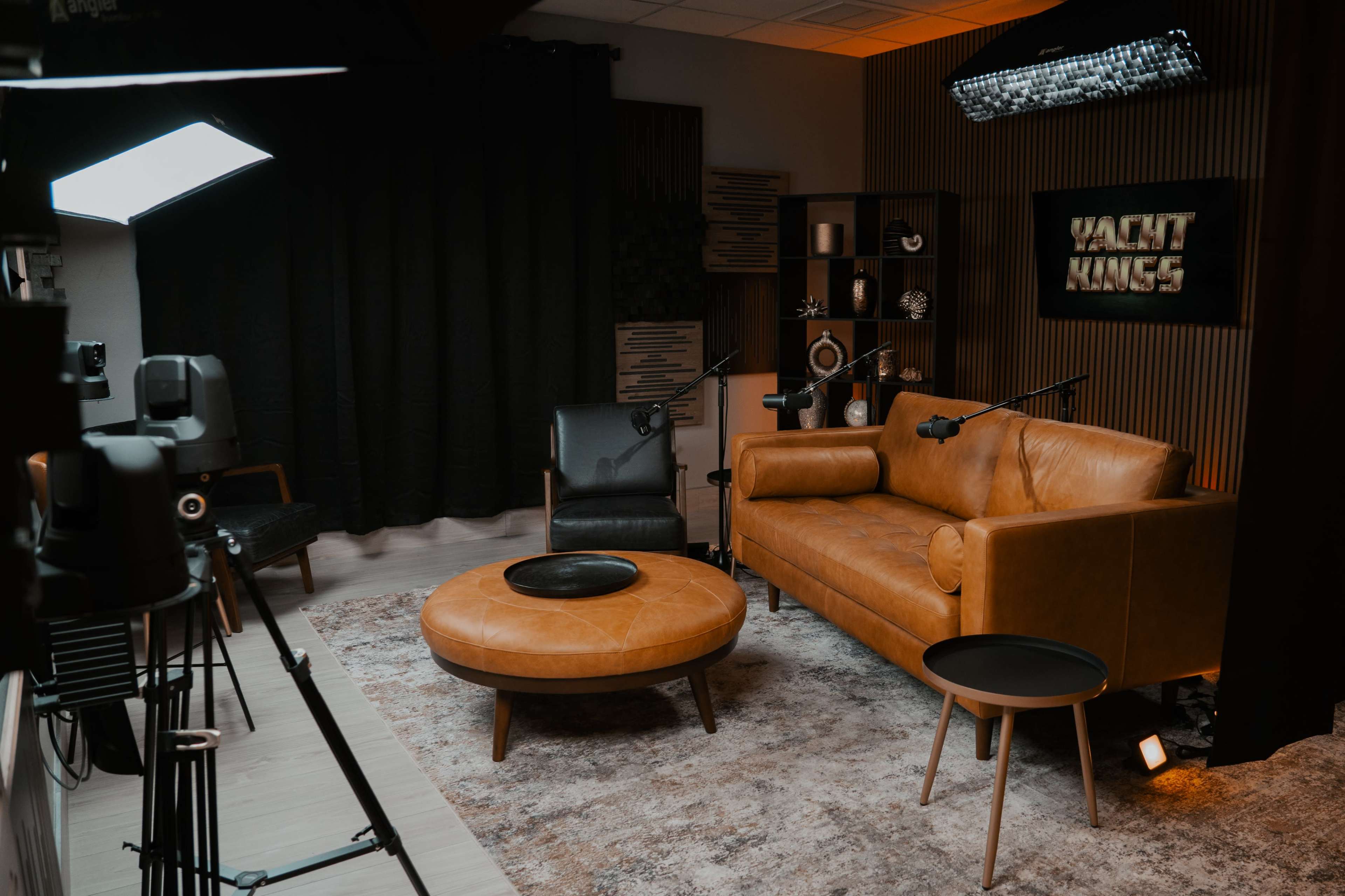 The image shows a studio setup with a brown leather sofa, a round coffee table, and two microphones positioned in front of two chairs, surrounded by various light sources and decorative elements.