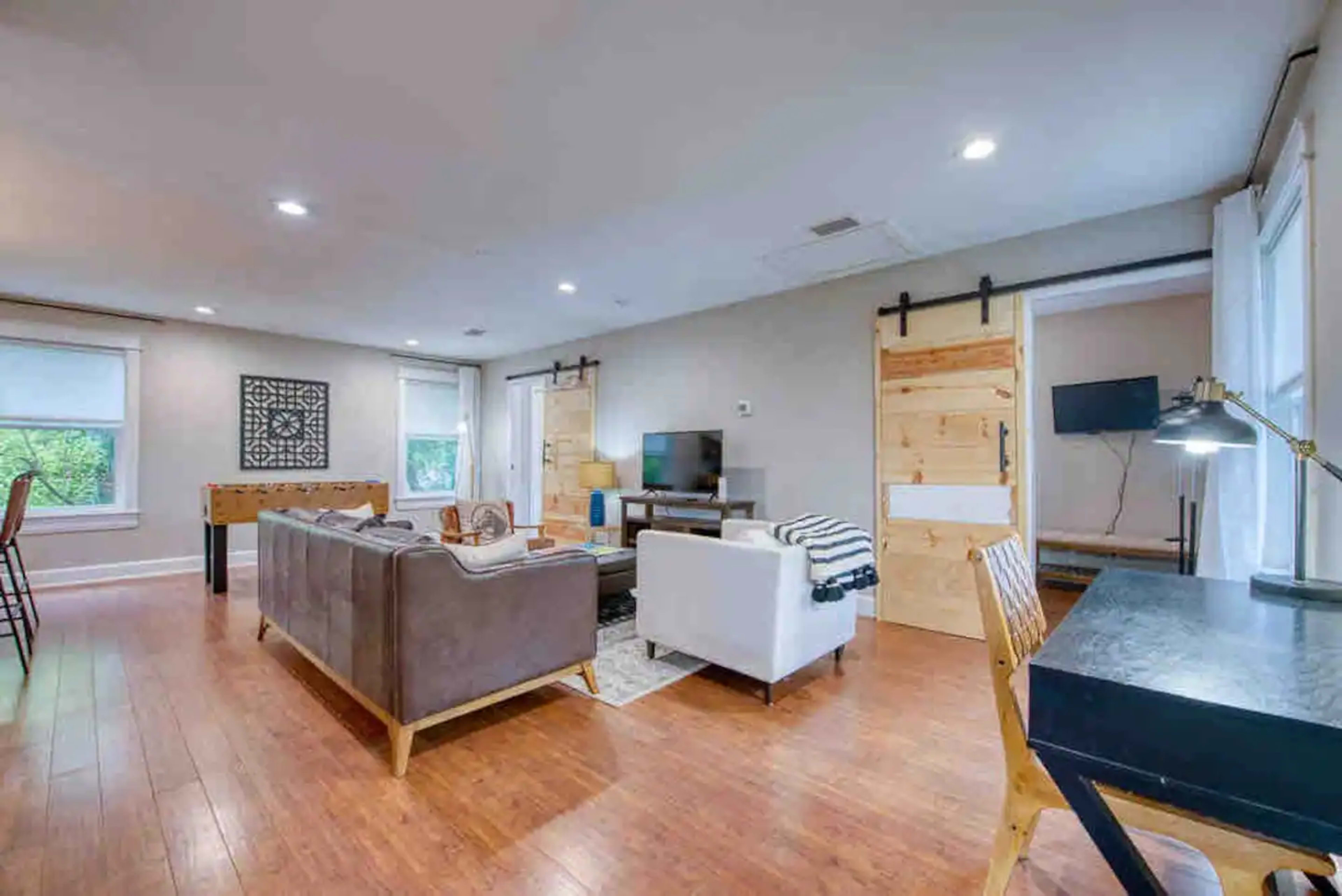 A modern living room with a brown couch, white armchair, television, and a desk, featuring wooden floors and sliding barn doors leading to another room.