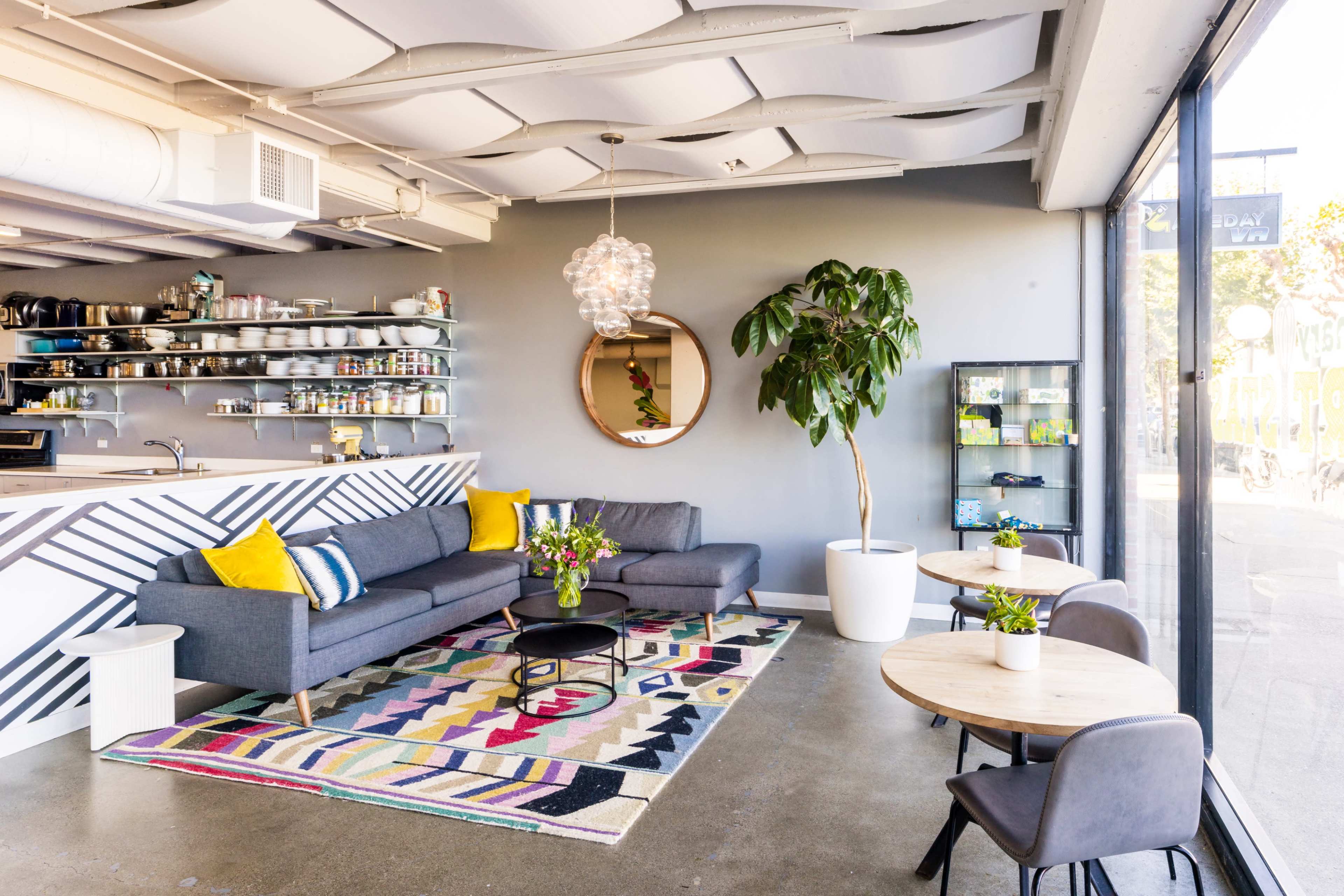 A modern café features a gray sofa, colorful patterned rug, round tables, and a large window with views of the outside.