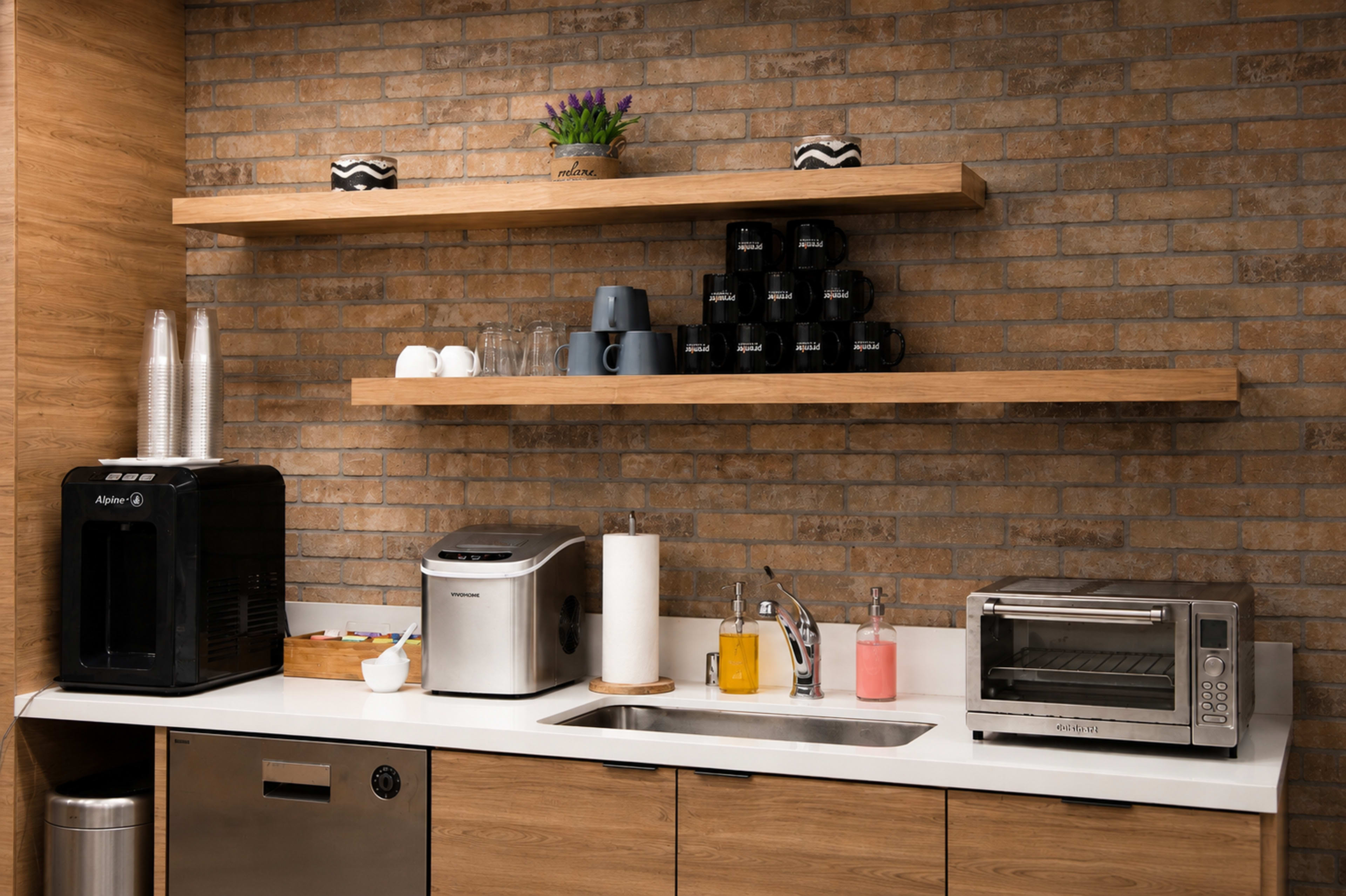 The image shows a modern kitchenette with wooden shelves displaying cups, a coffee maker, a toaster, and various containers, along with a sink and countertop.