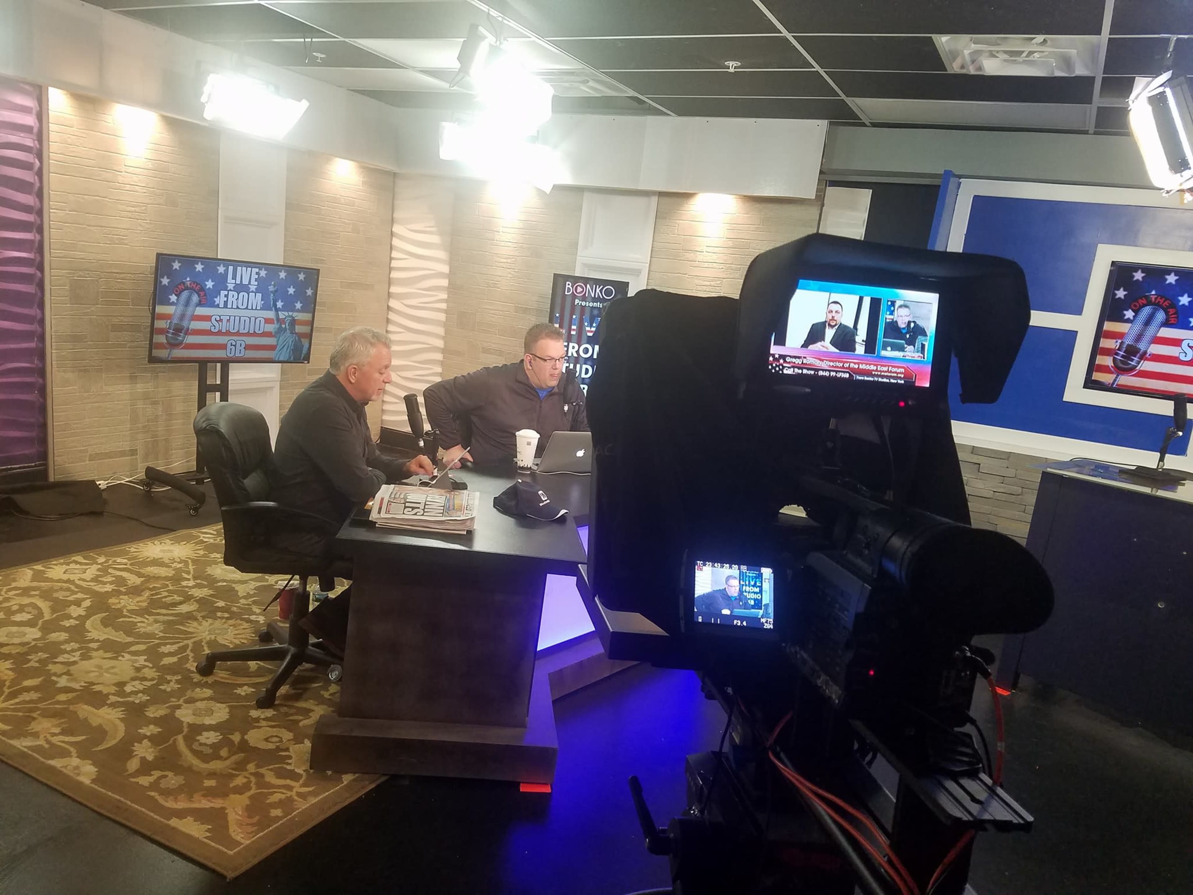 Two men sit at a desk with a laptop in a television studio, while a camera captures the scene in the foreground.