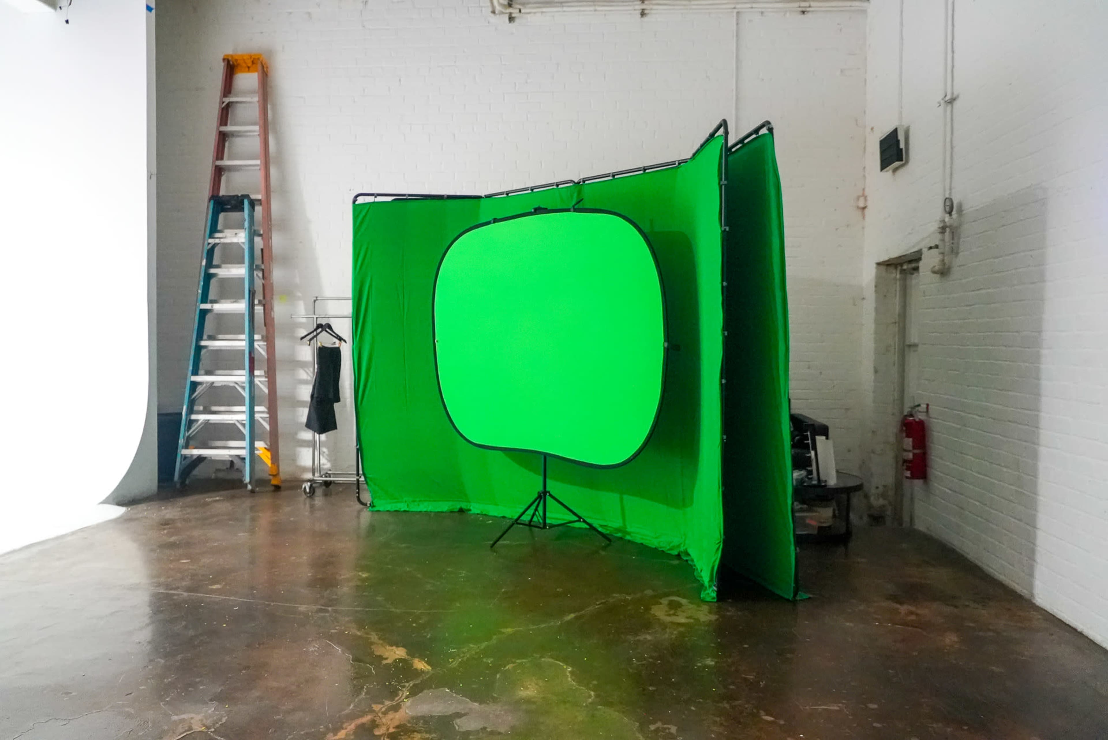 A green screen setup is positioned in a studio space with a ladder and clothing rack nearby.