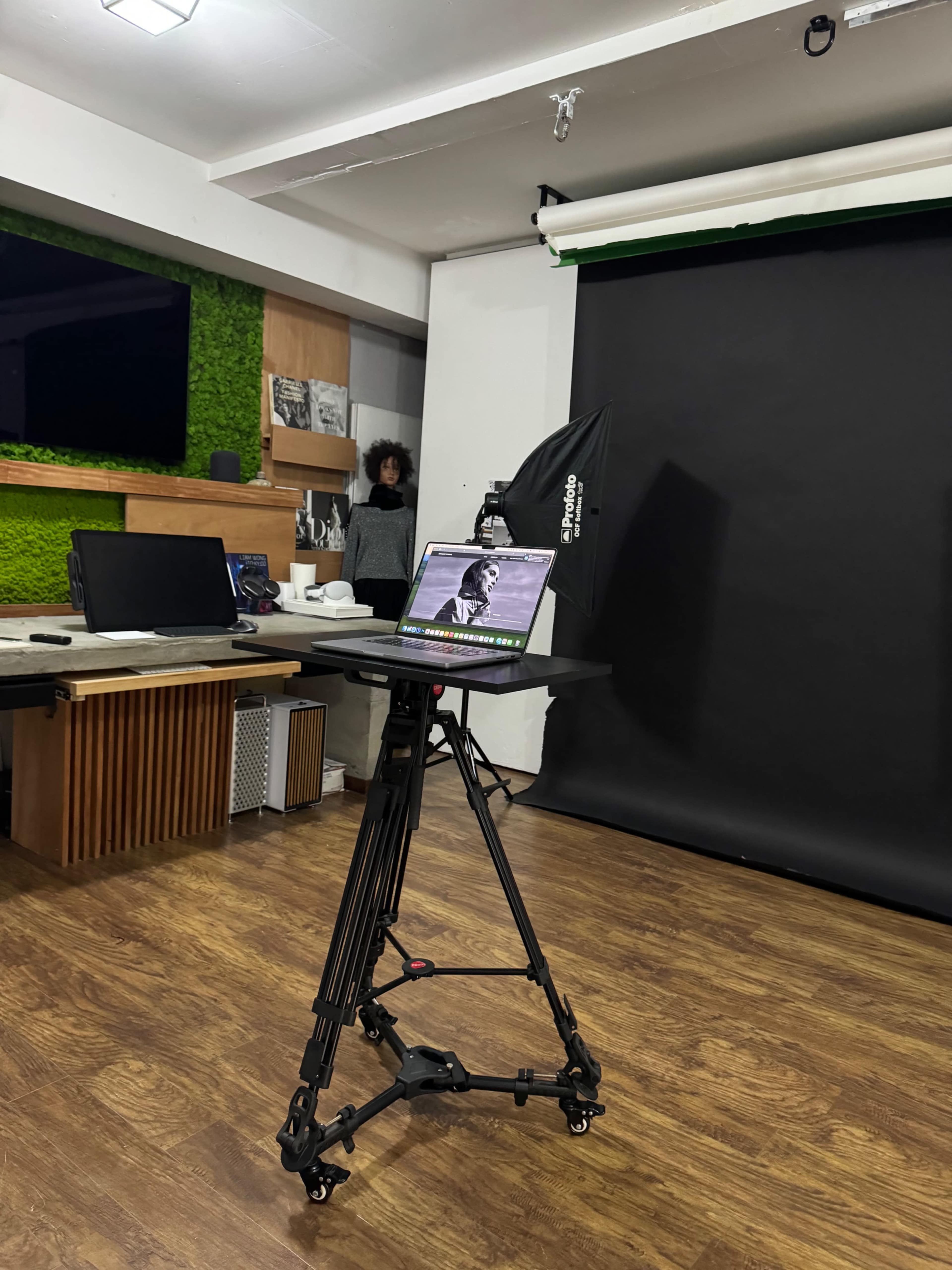 A laptop on a movable stand is set up in a photography studio with a black backdrop and green accents in the decor.