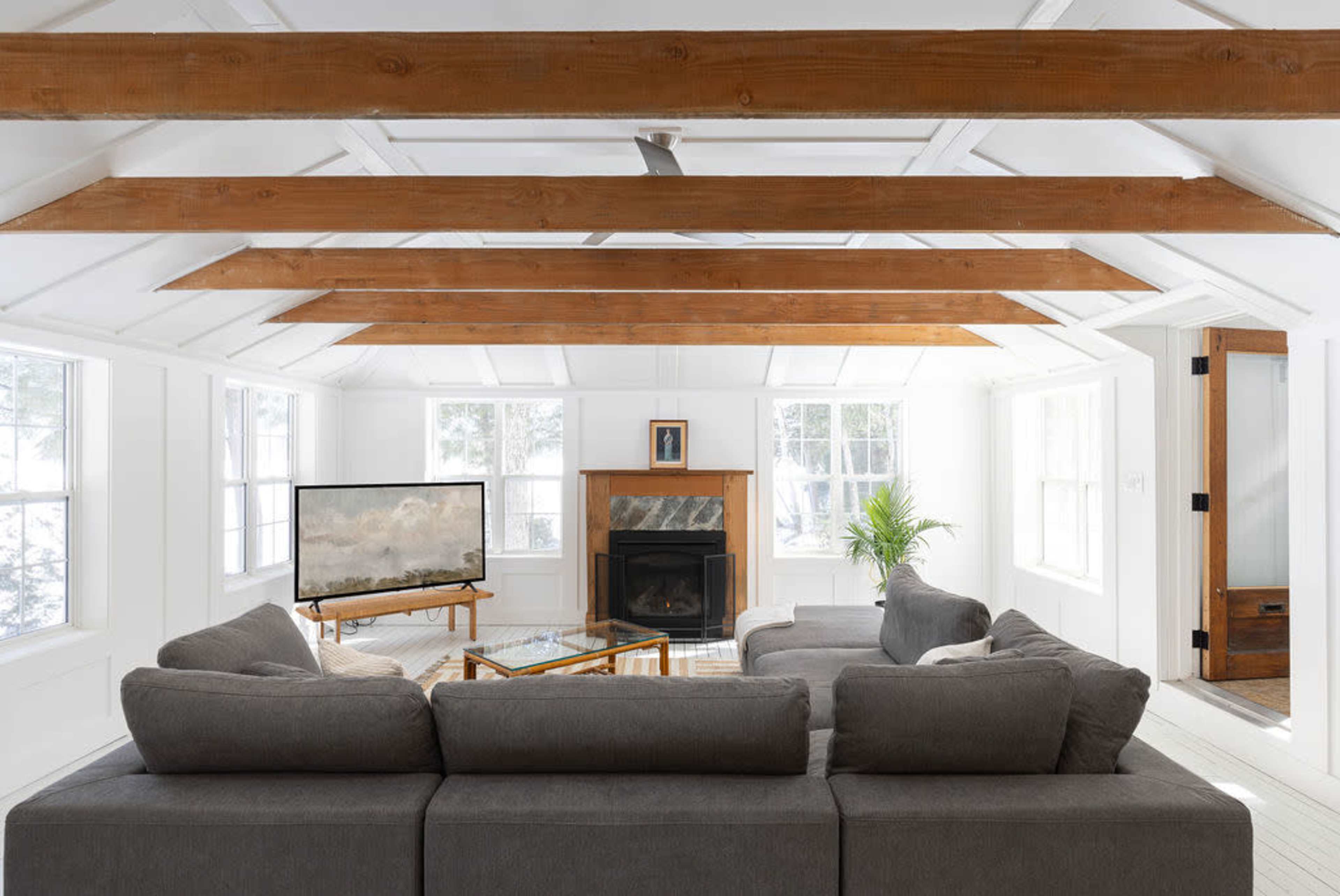 The image shows a bright, modern living room with a gray sectional sofa, a television, a fireplace, and wooden beams on the ceiling.