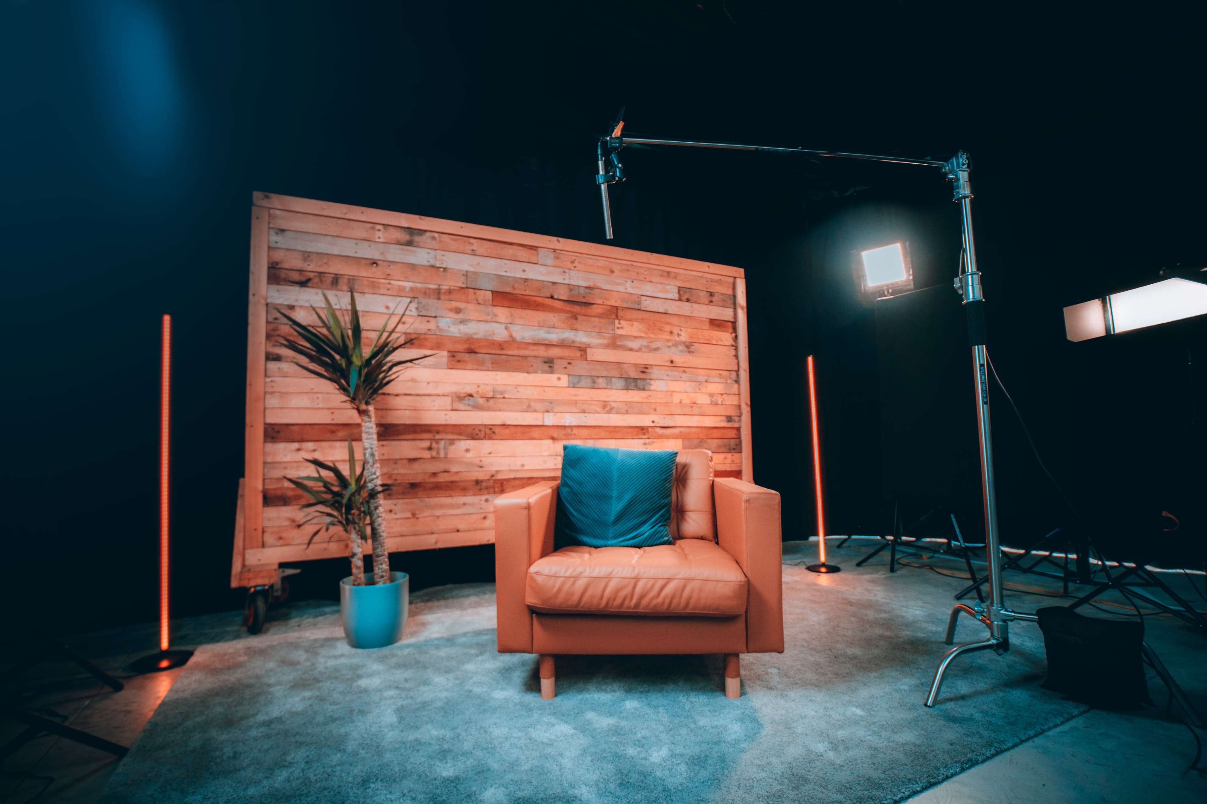 A modern video setup featuring a brown leather armchair in front of a wooden panel backdrop, with studio lights and a potted plant nearby.