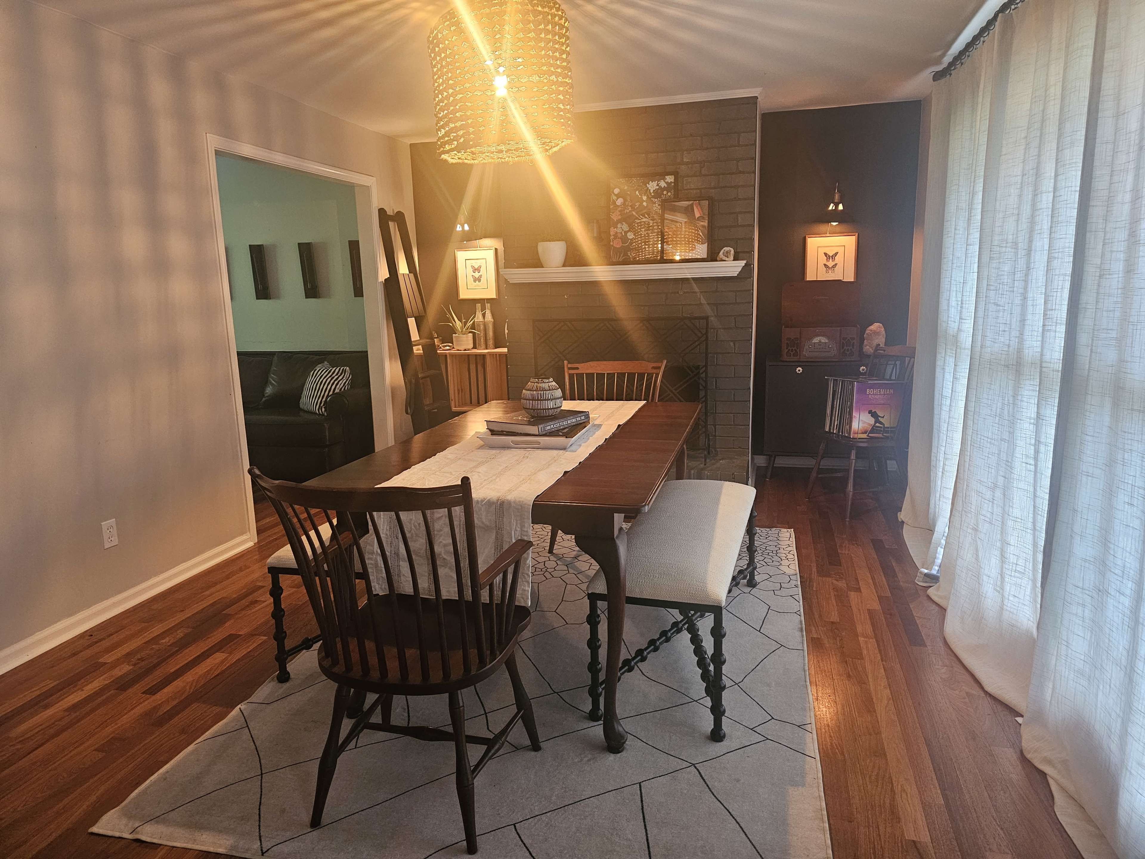 The image shows a dining area featuring a wooden table with chairs, a patterned rug, a fireplace, and large windows draped with light-colored curtains.