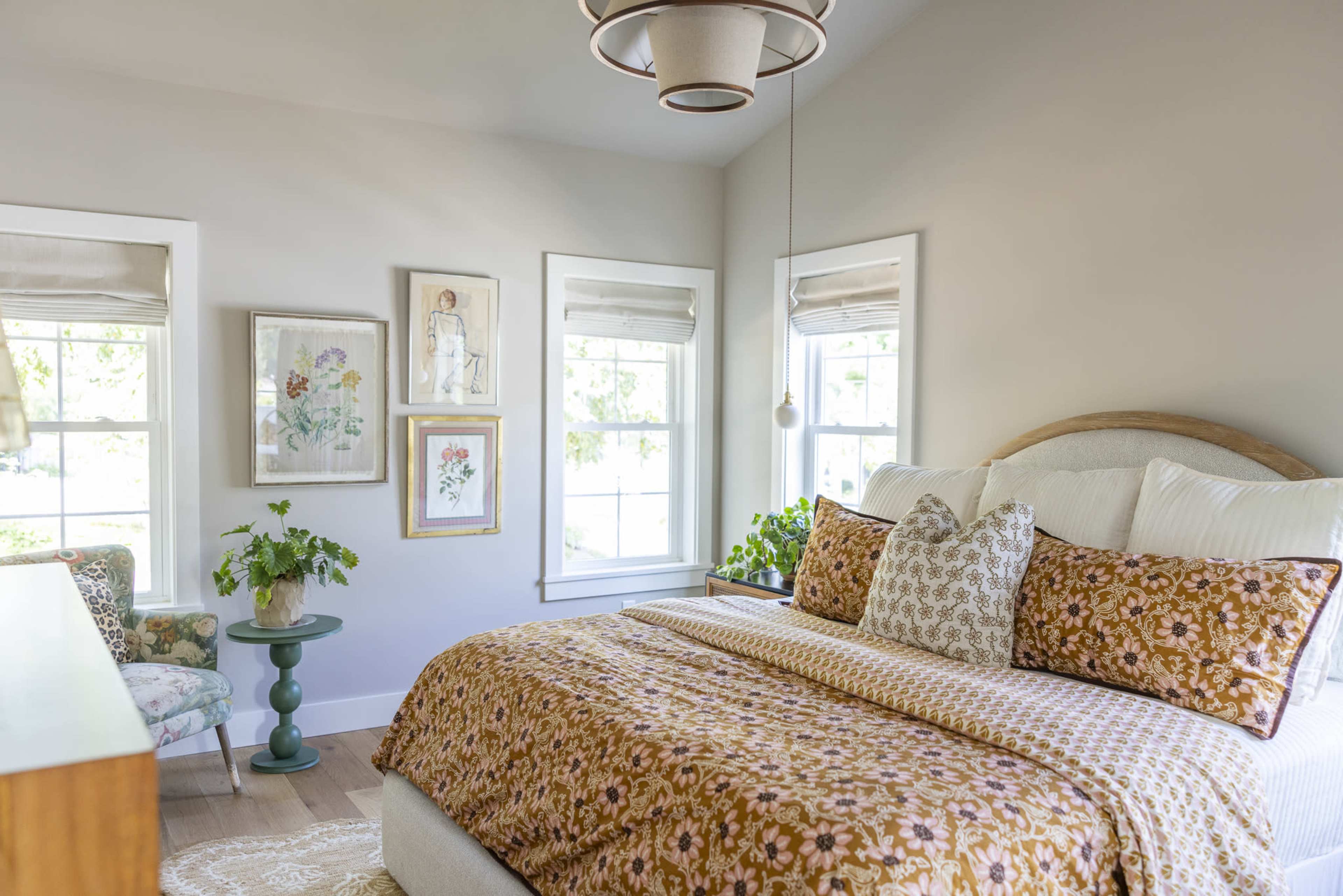 A bright bedroom features a bed with a patterned brown and white comforter, surrounded by windows and framed artwork on the walls.