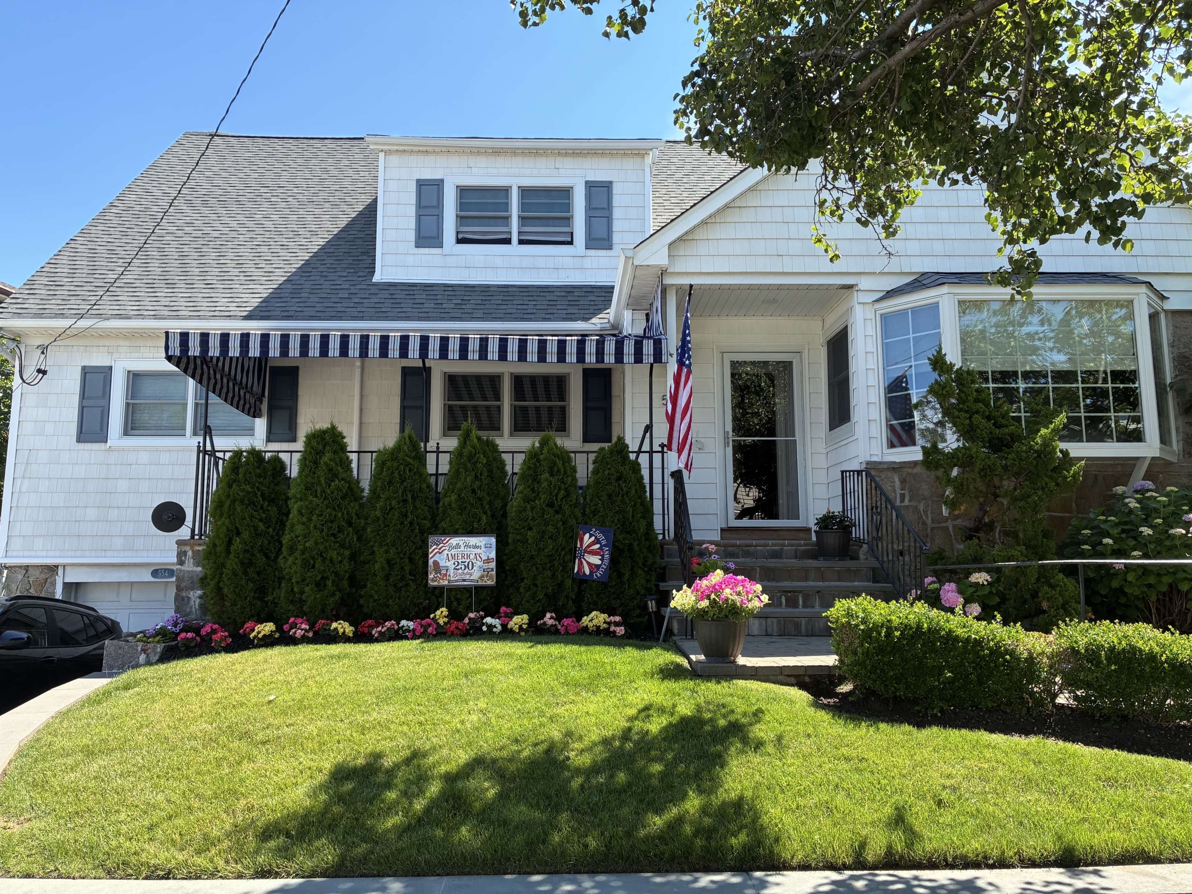 A residential home features a front yard with neatly manicured grass, vibrant flower beds, and an American flag displayed on the porch.