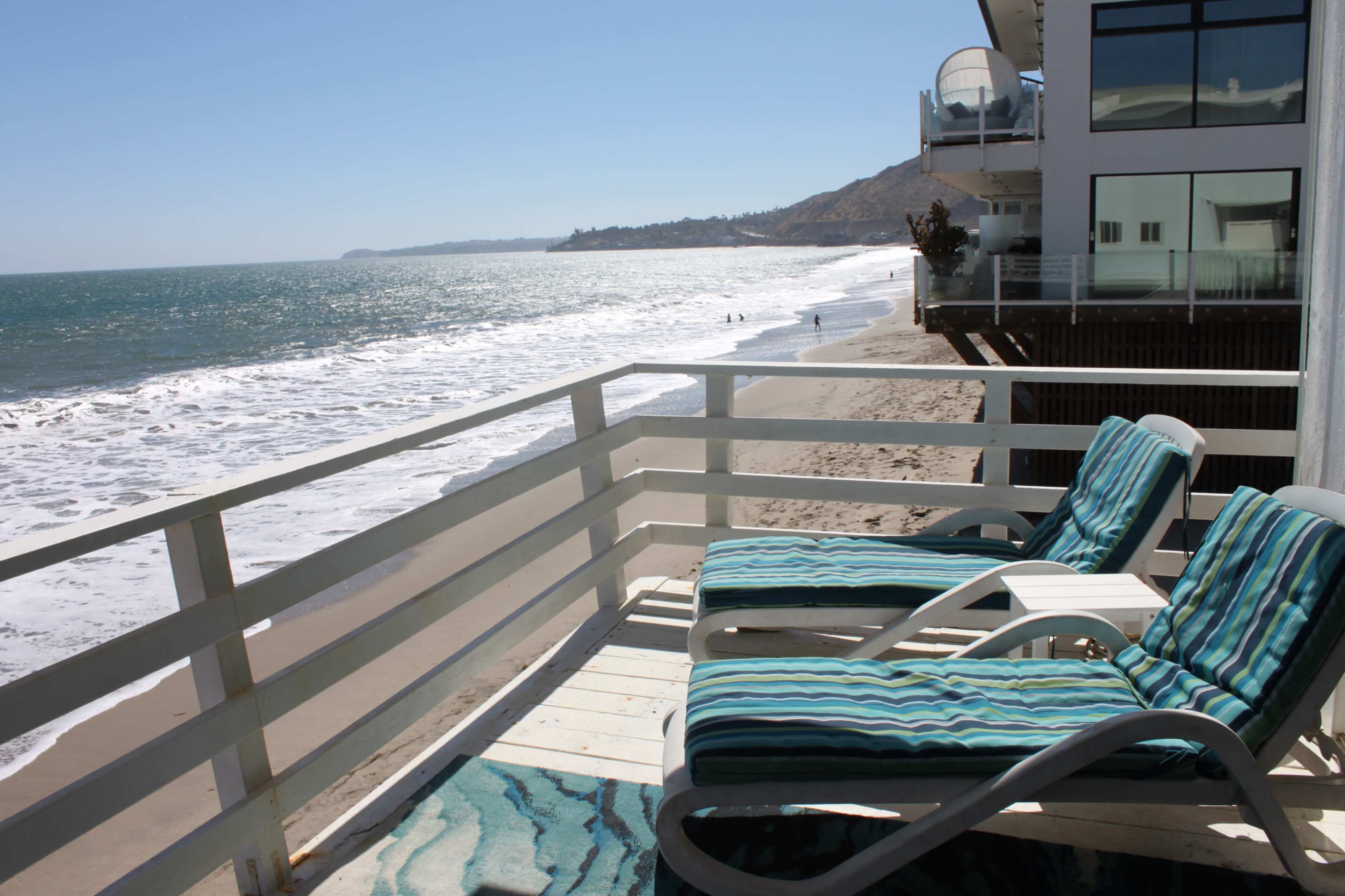 A balcony with two striped chaise lounges overlooks a sandy beach and the ocean waves.