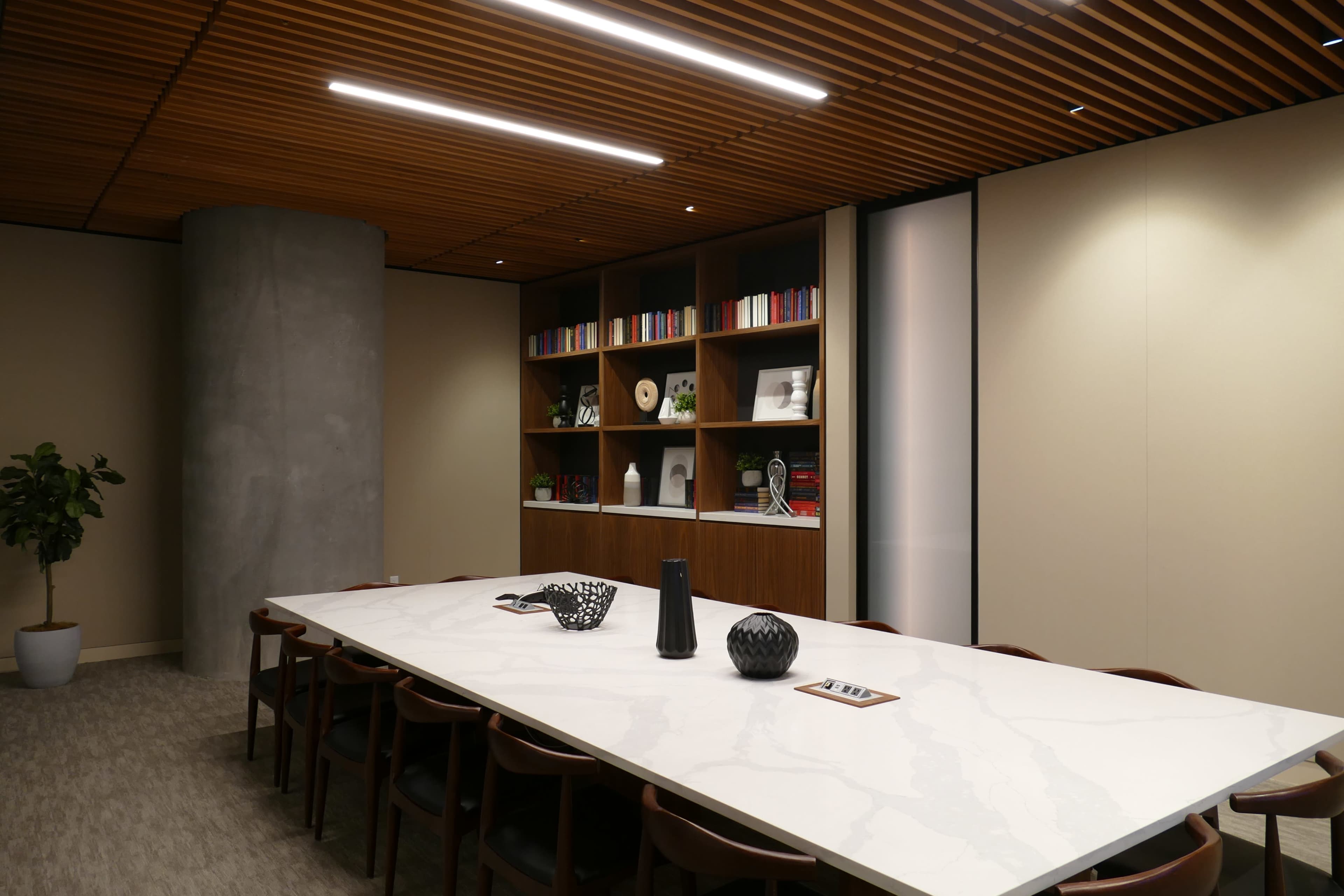 The image shows a modern conference room featuring a large marble table, wooden ceiling panels, and built-in shelves with decorative items and books.