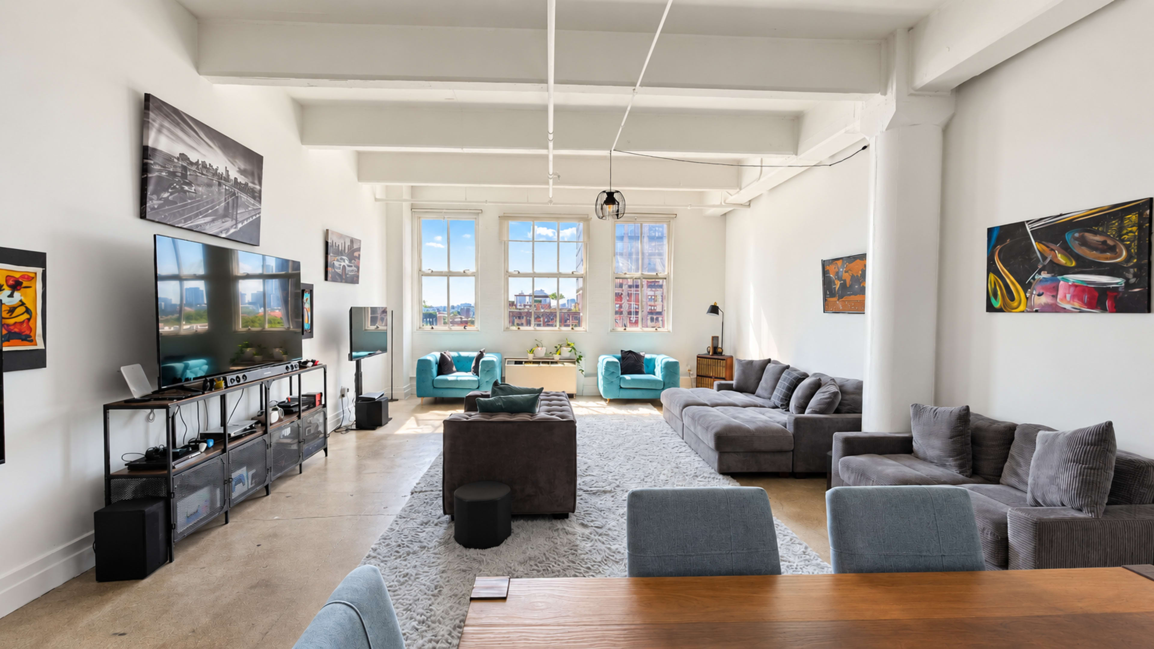 The image depicts a spacious and well-lit living room with modern furnishings, including a large TV, multiple sofas, and windows offering a view of the outdoors.