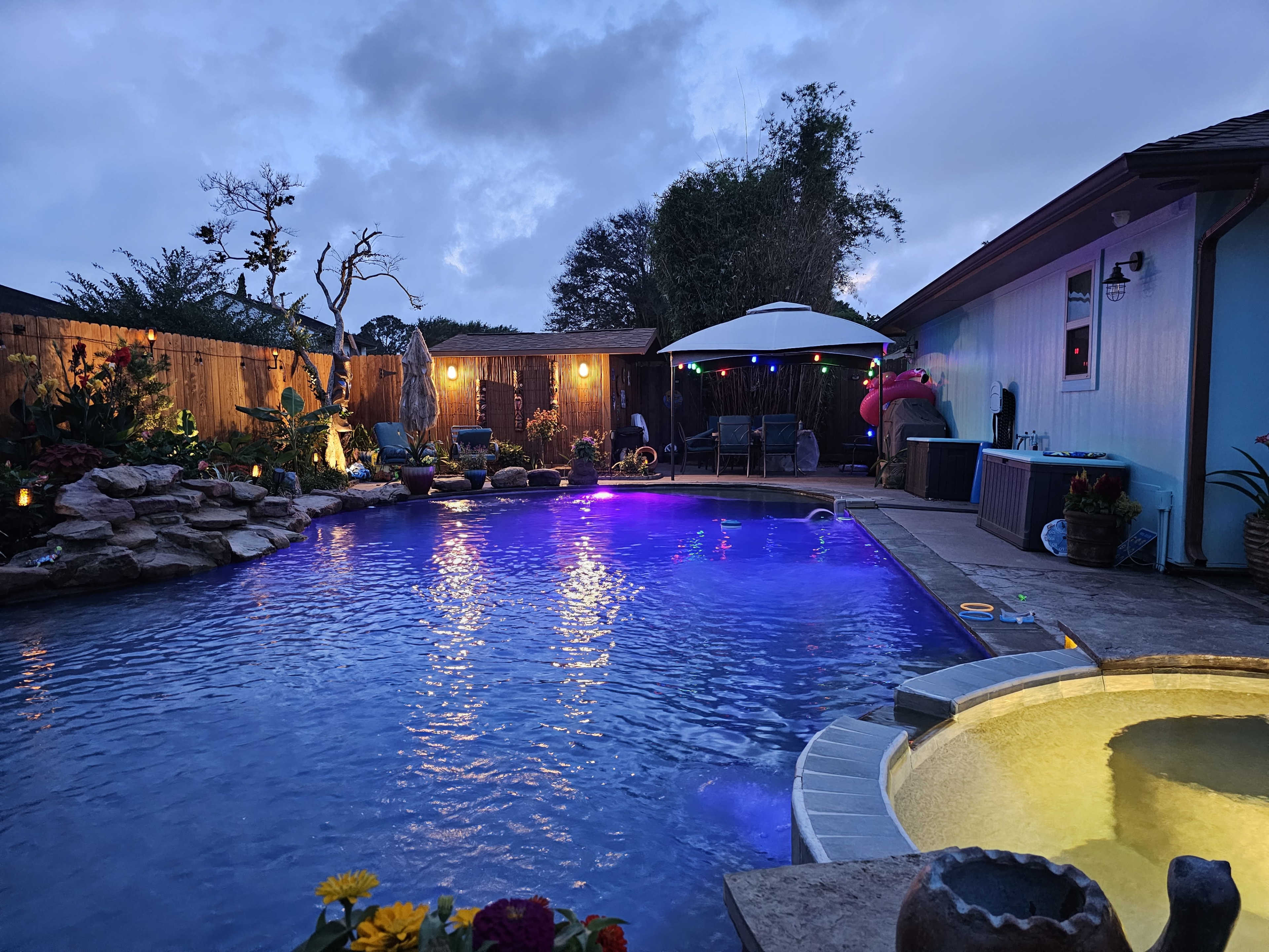 The image shows a backyard pool area illuminated with purple lights, featuring a hot tub and surrounded by tropical plants and a shaded seating area.