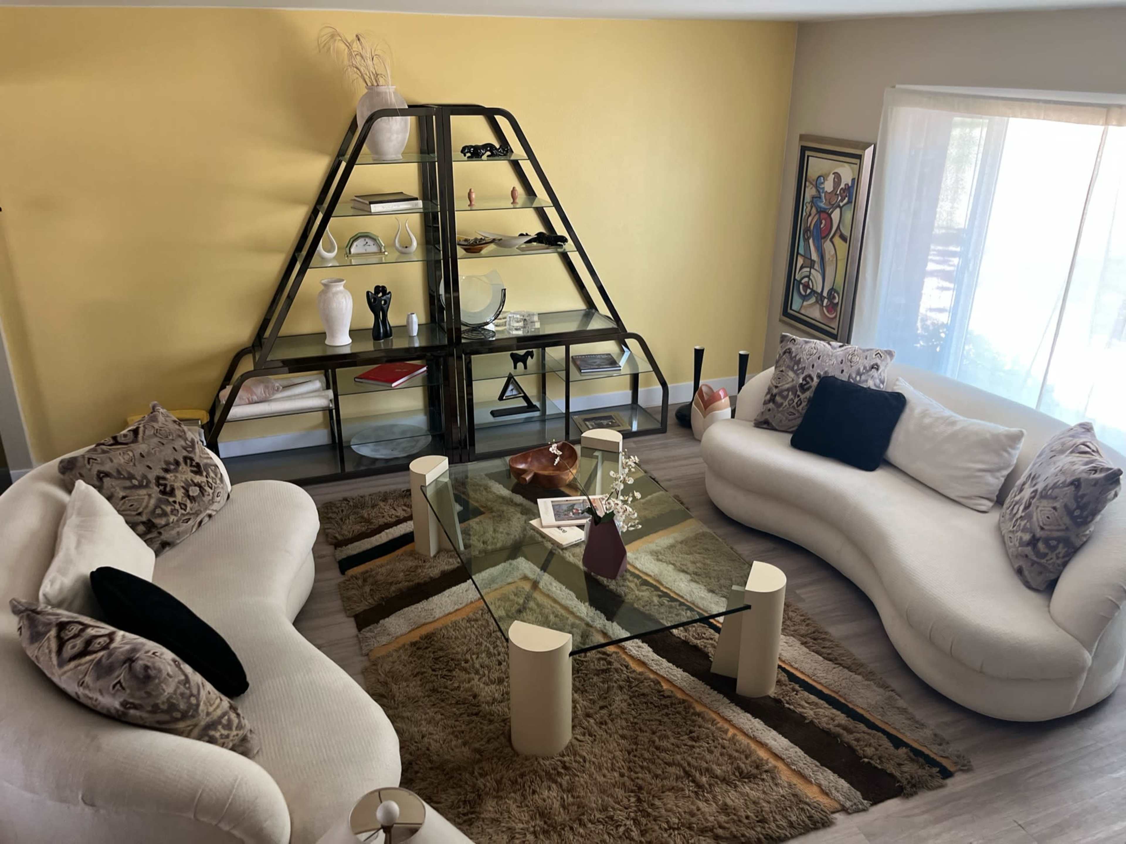A living room with two curved white sofas facing a glass coffee table on a patterned rug, surrounded by a yellow wall and a set of shelves displaying various decor items.