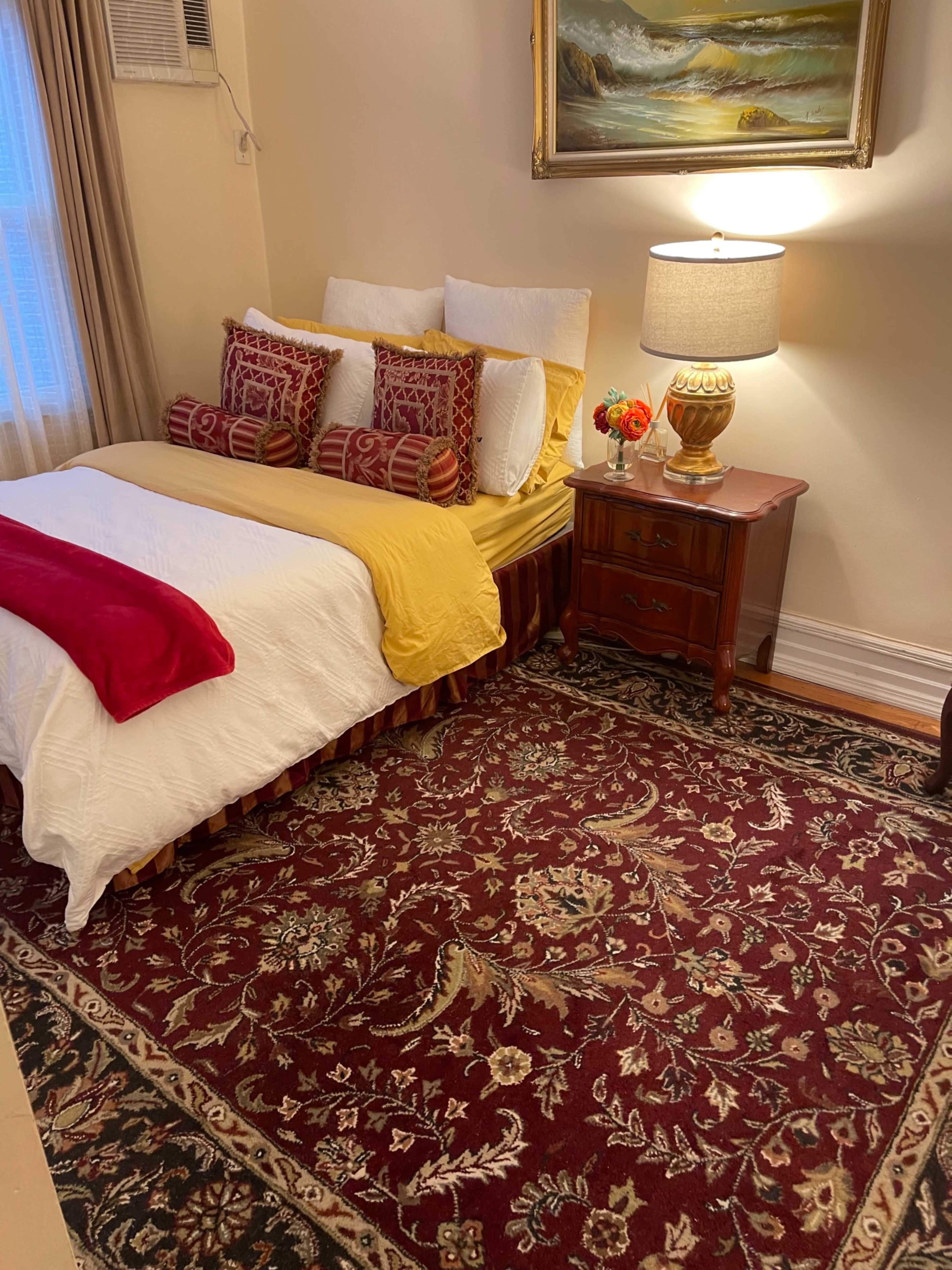 A neatly arranged bedroom featuring a bed with yellow and white bedding, decorative pillows, a nightstand with a lamp and flowers, and a large area rug.