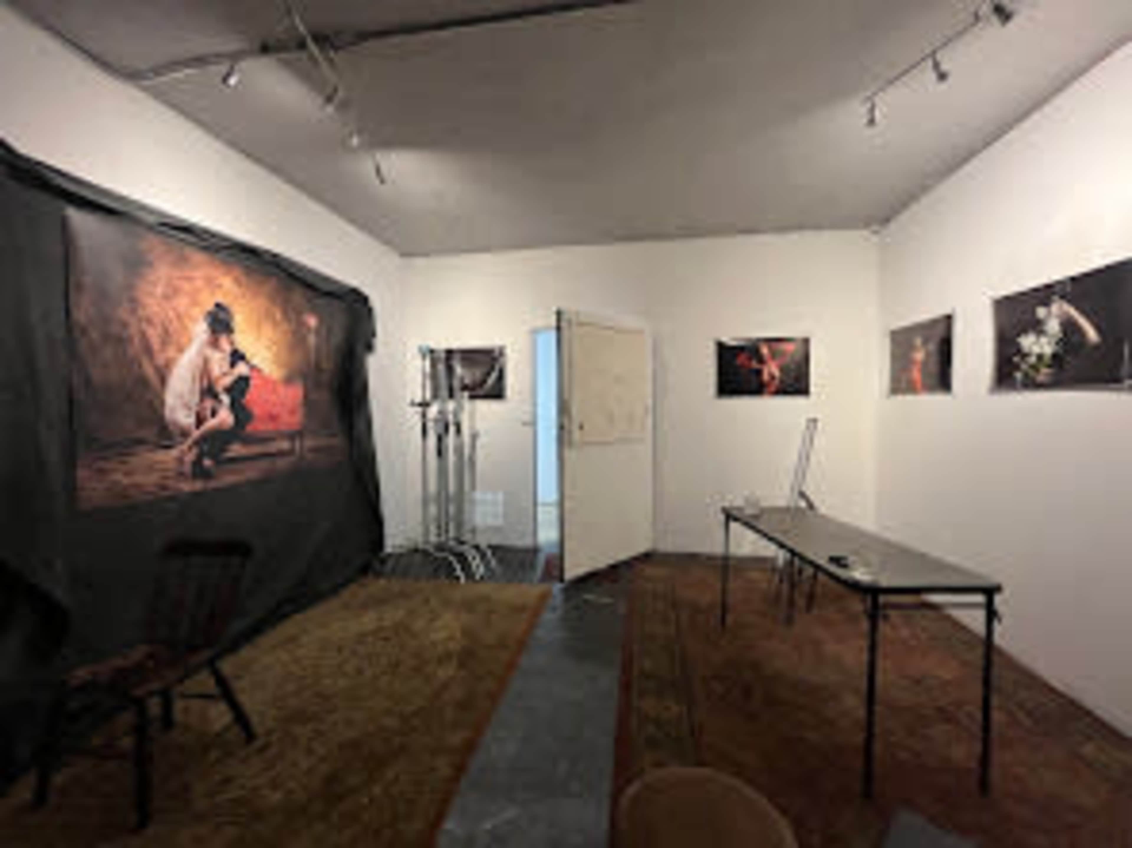 The image shows an indoor art studio featuring a black backdrop, several framed photographs on the walls, and a table with a chair.