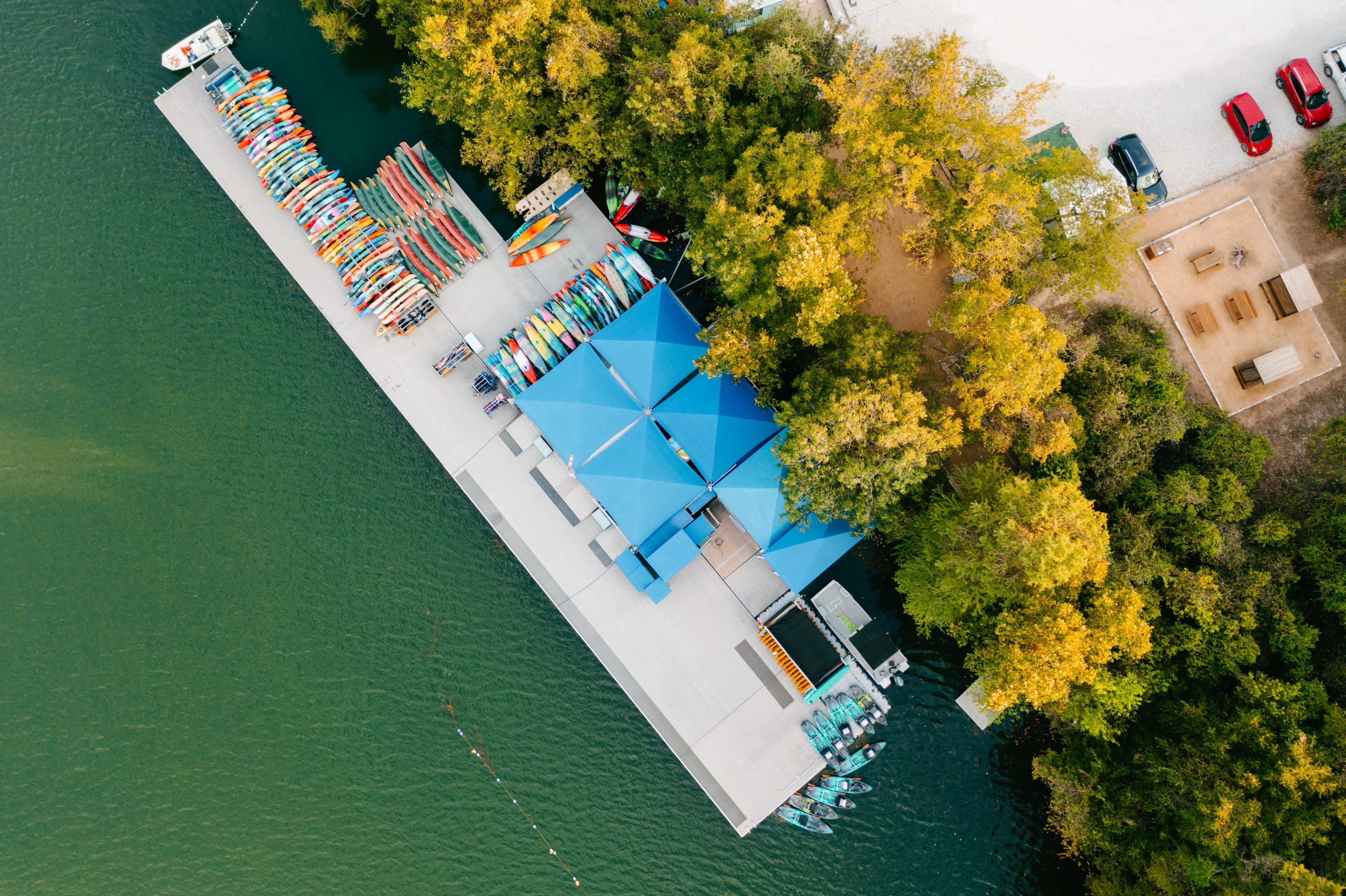 An aerial view shows a dock with colorful kayaks and canoes lined up, surrounded by trees and a small recreational area with picnic tables.