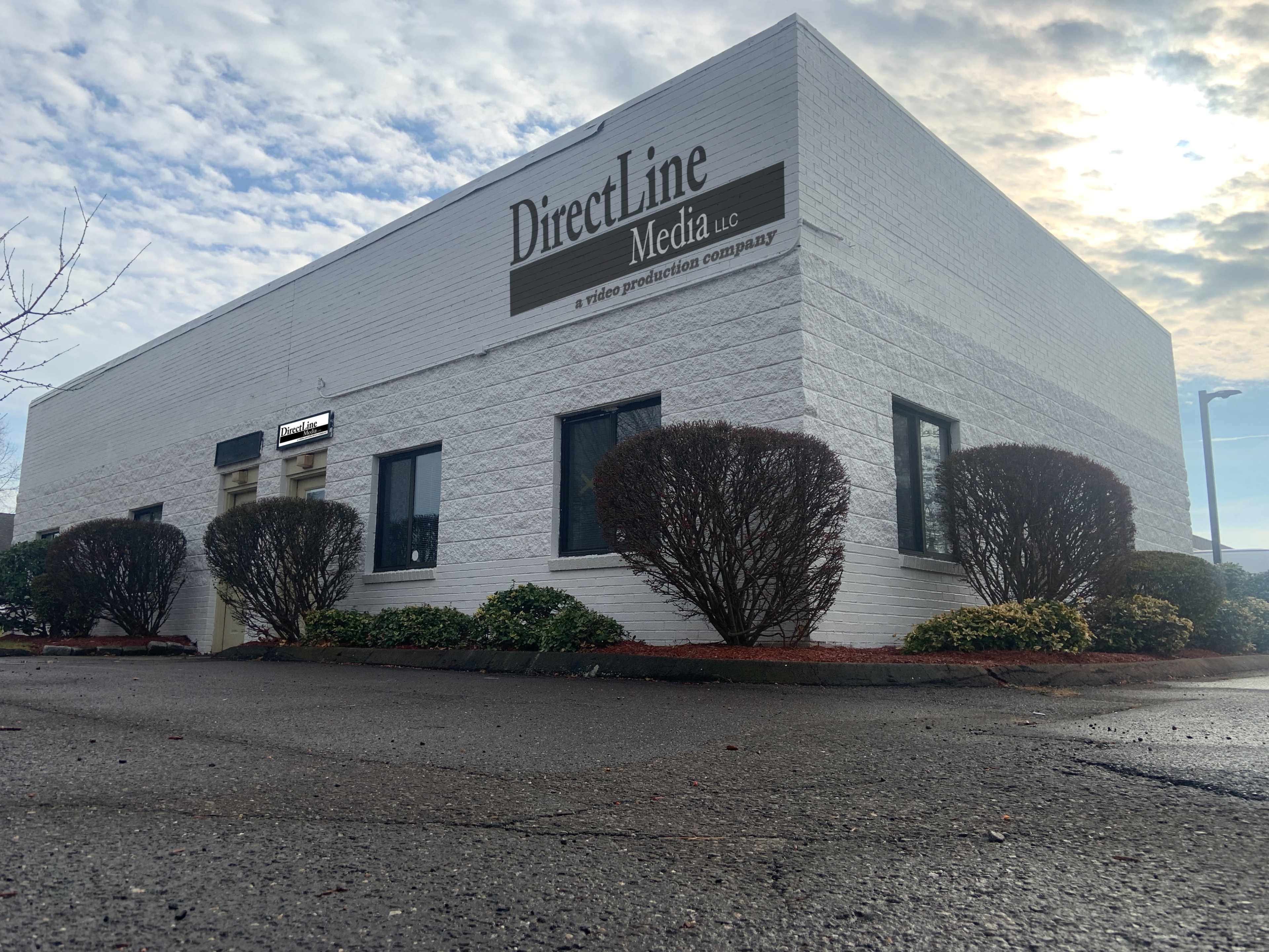A one-story white building with "DirectLine Media" signage and landscaping, situated on a paved area under a cloudy sky.