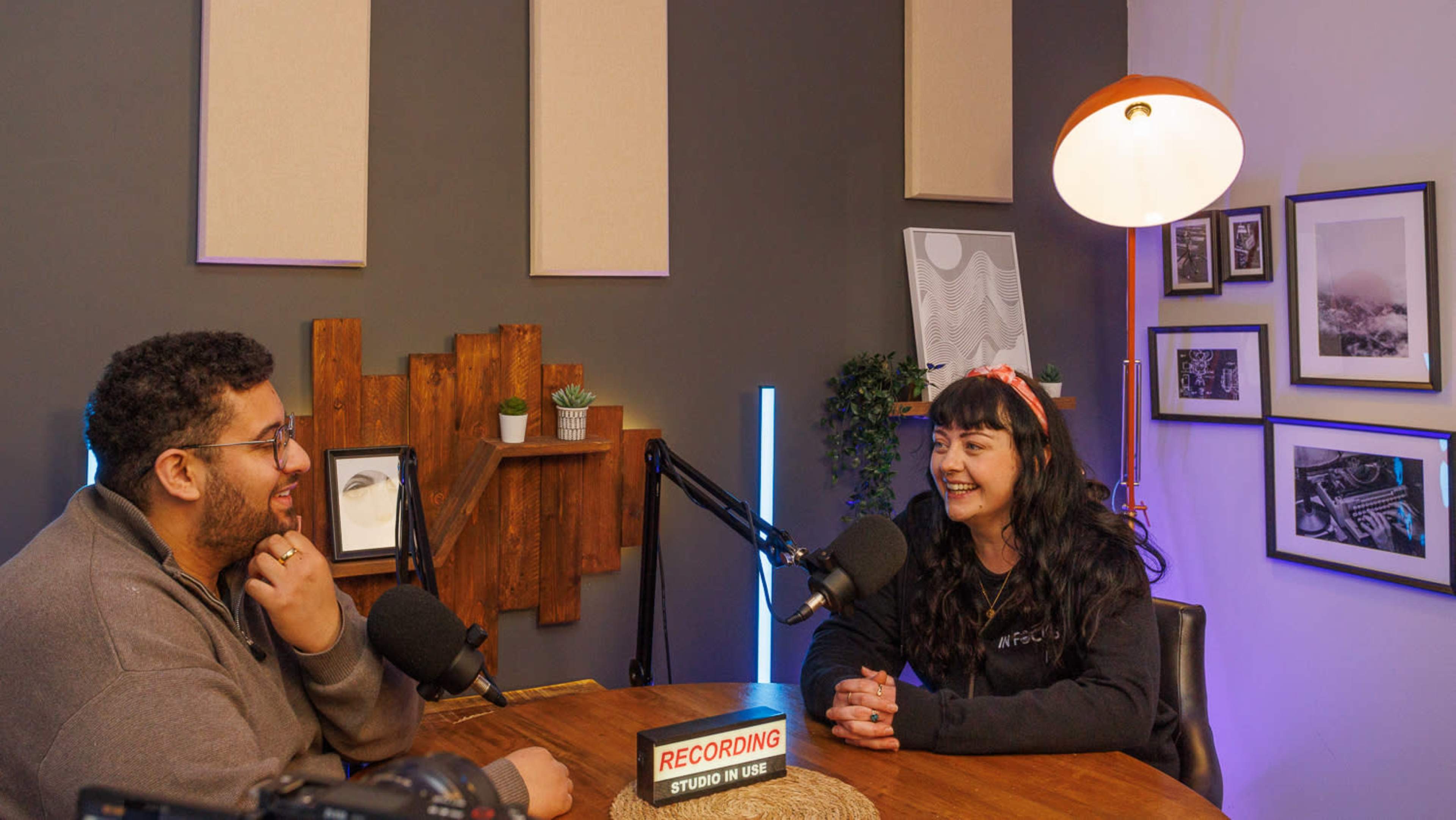 Two people are engaged in a conversation at a podcast recording studio, seated at a table with microphones in front of them and decorative wall elements in the background.