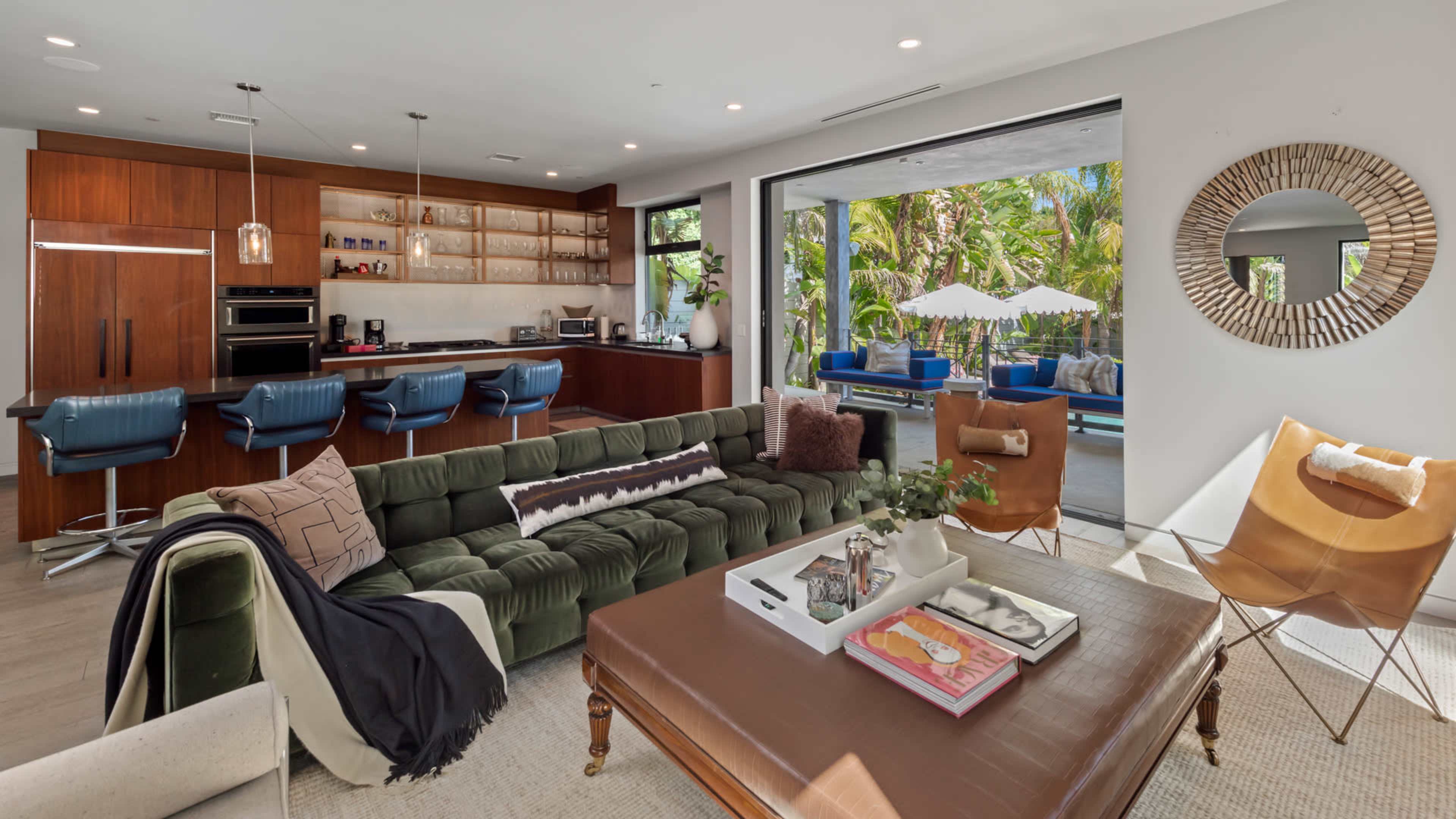 A modern living space features a green sectional sofa, a wooden coffee table, and a kitchen with bar seating, surrounded by large windows revealing a patio area with outdoor seating.