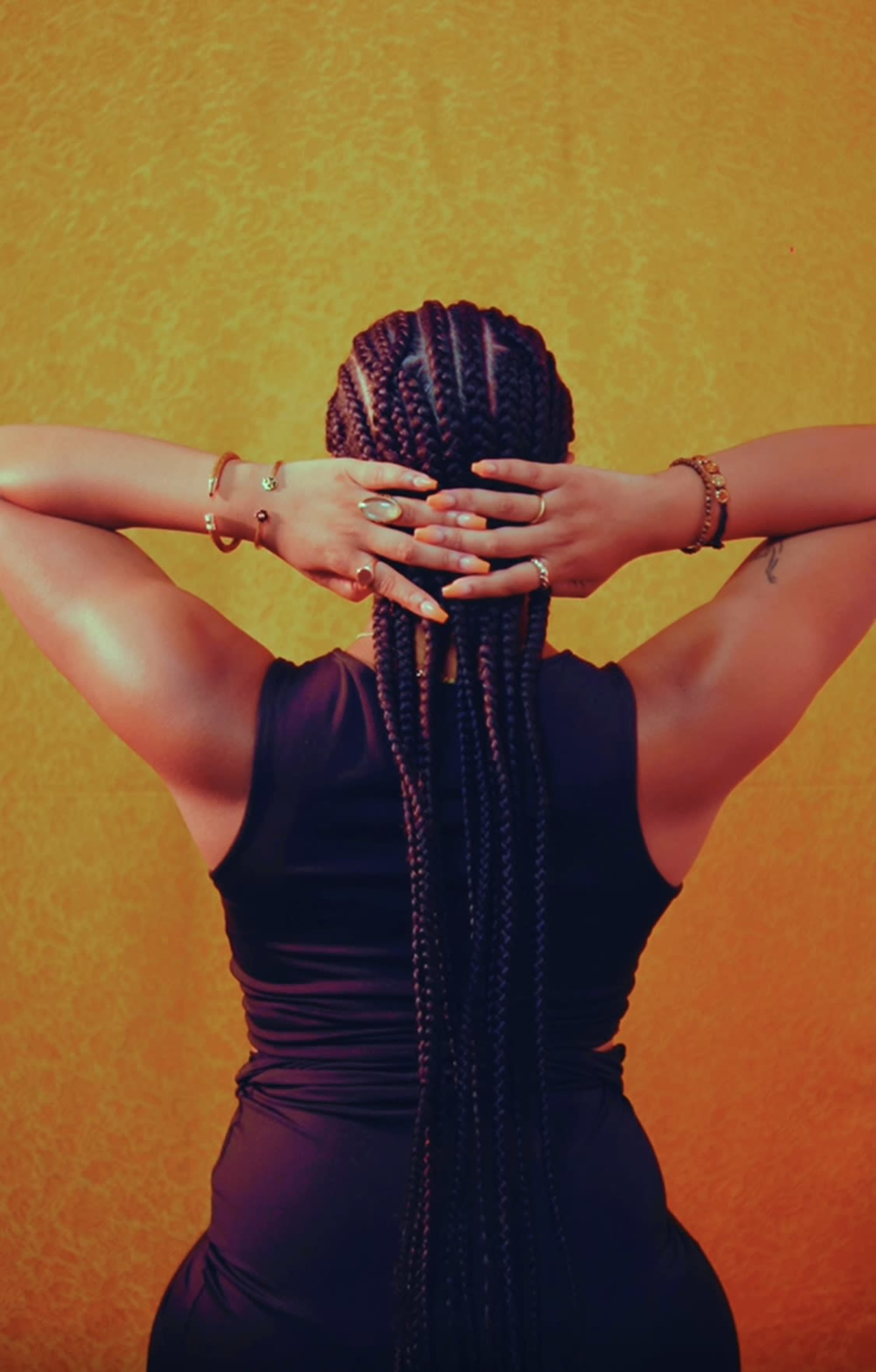 A person with long braided hair stands with their back to the camera, holding their hair up against a bright yellow background.