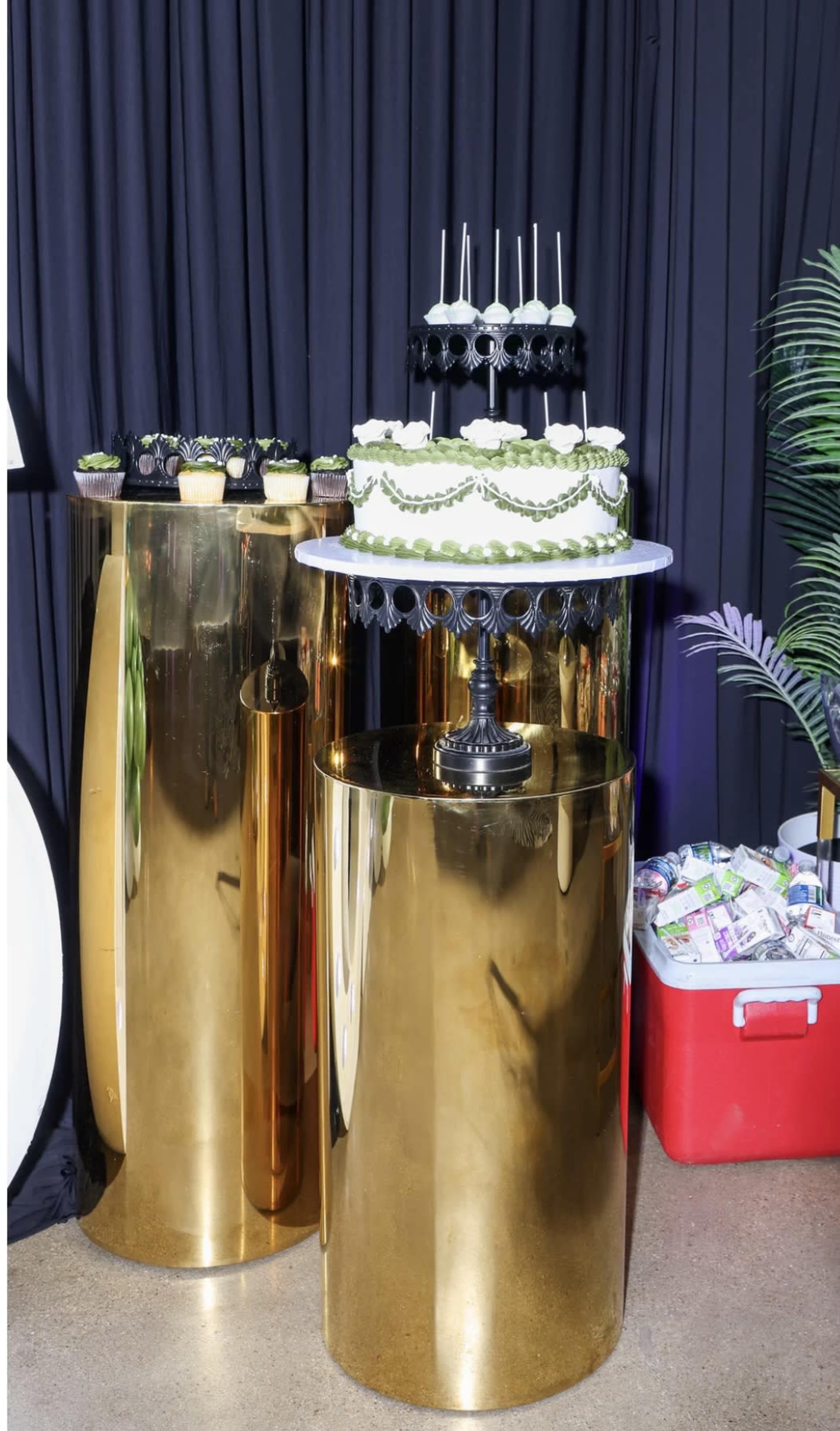 The image shows two shiny gold tables, one displaying a multi-tiered cake decorated with green accents and the other with an assortment of cupcakes.