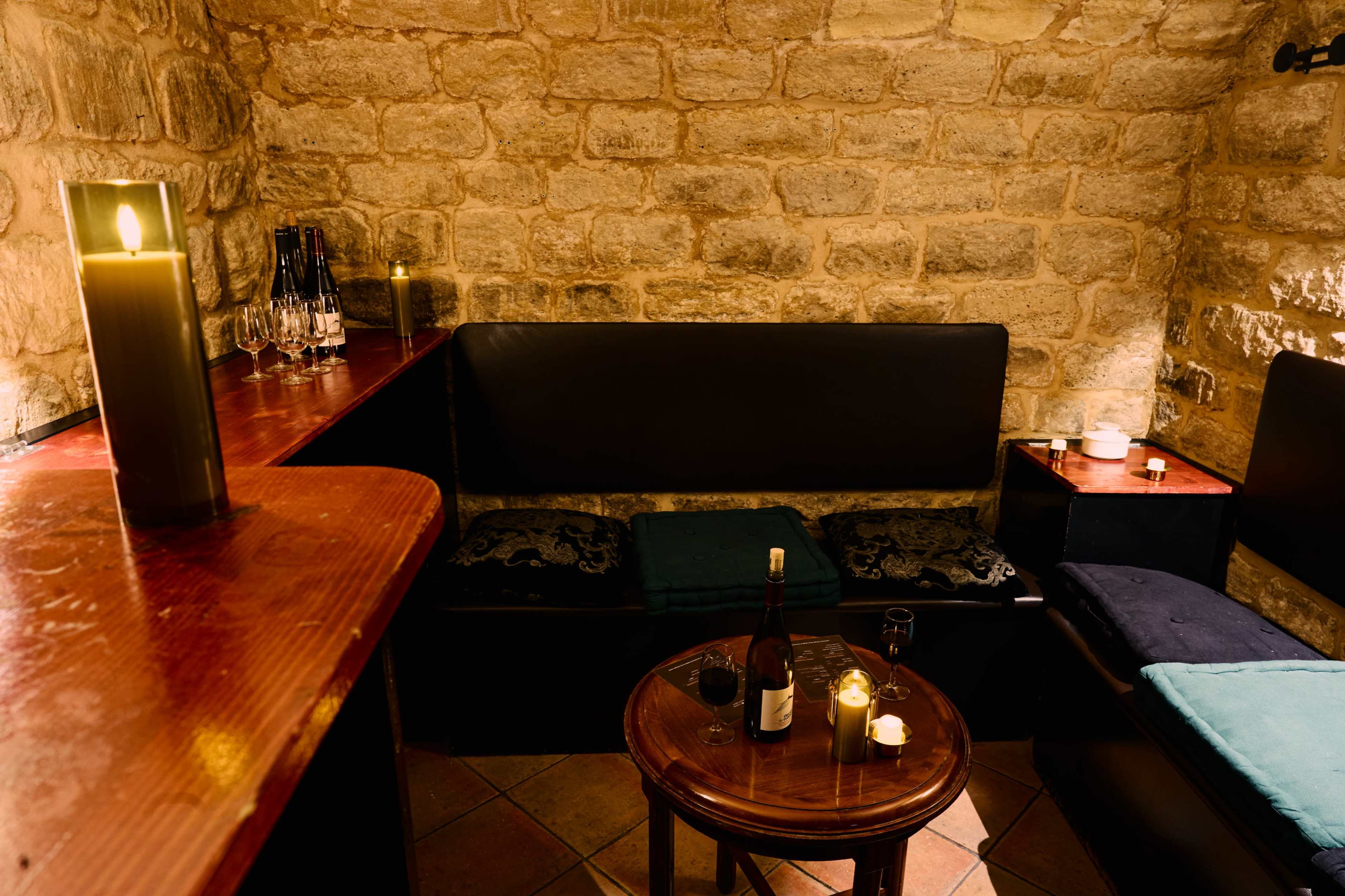 The image shows a cozy interior of a dimly lit room with stone walls, featuring a wooden table surrounded by black seating and a candle placed on the table.