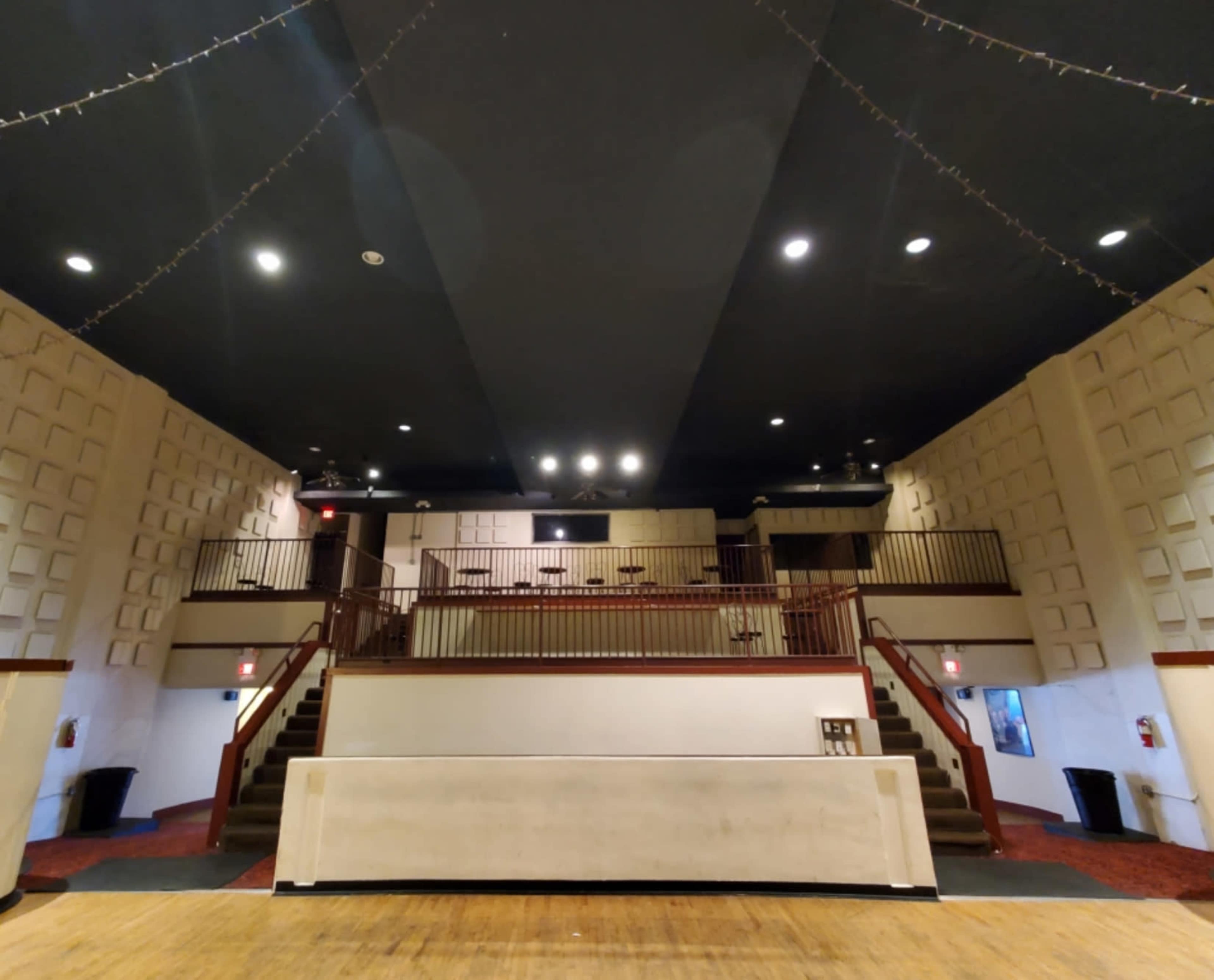 The image shows the interior of a theater, featuring a stage area, seating on a raised level, and a dark ceiling with lights hanging above.