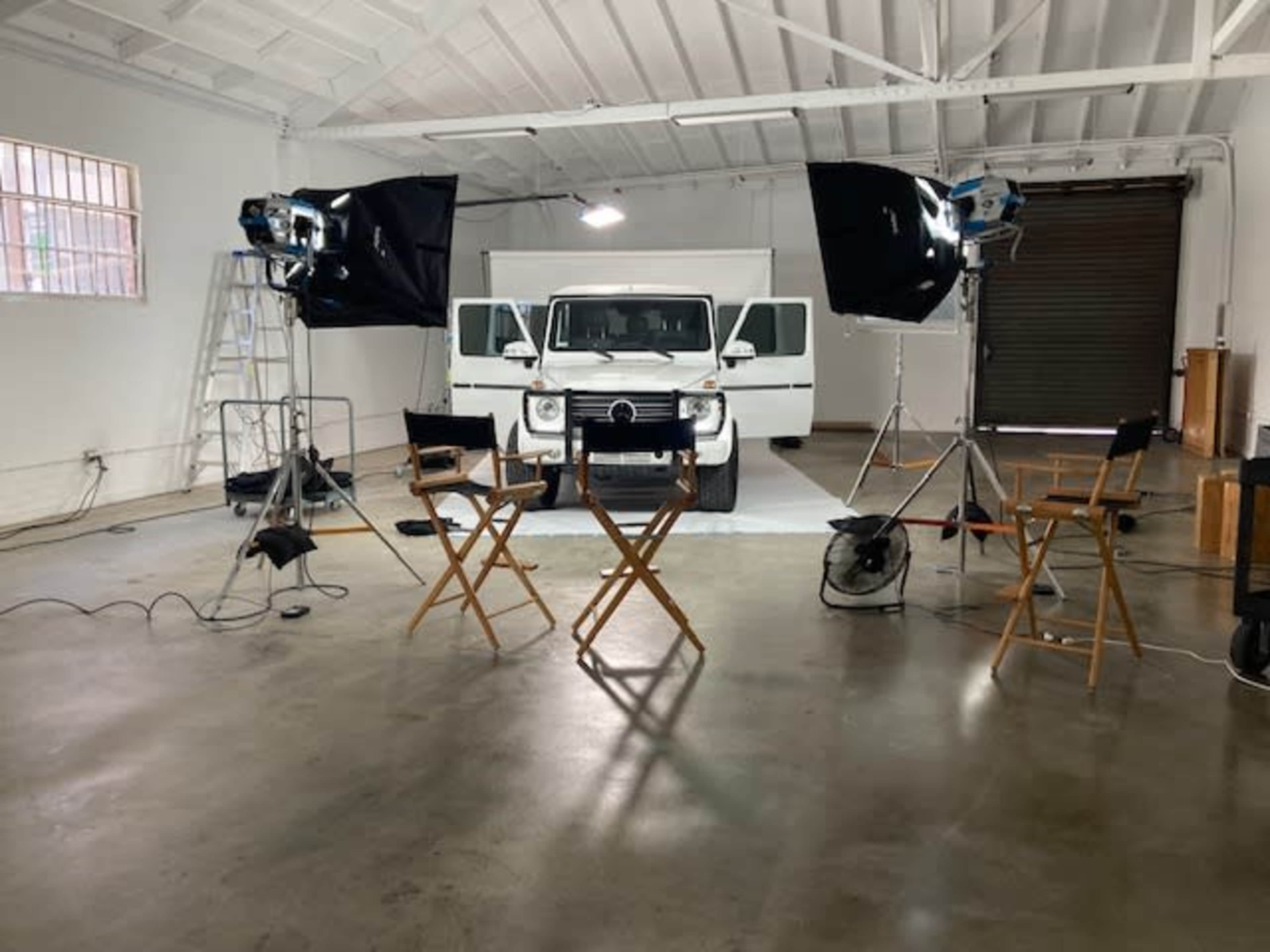 A studio setup with lights, cameras, and two director's chairs facing a white vehicle positioned in front of a backdrop.
