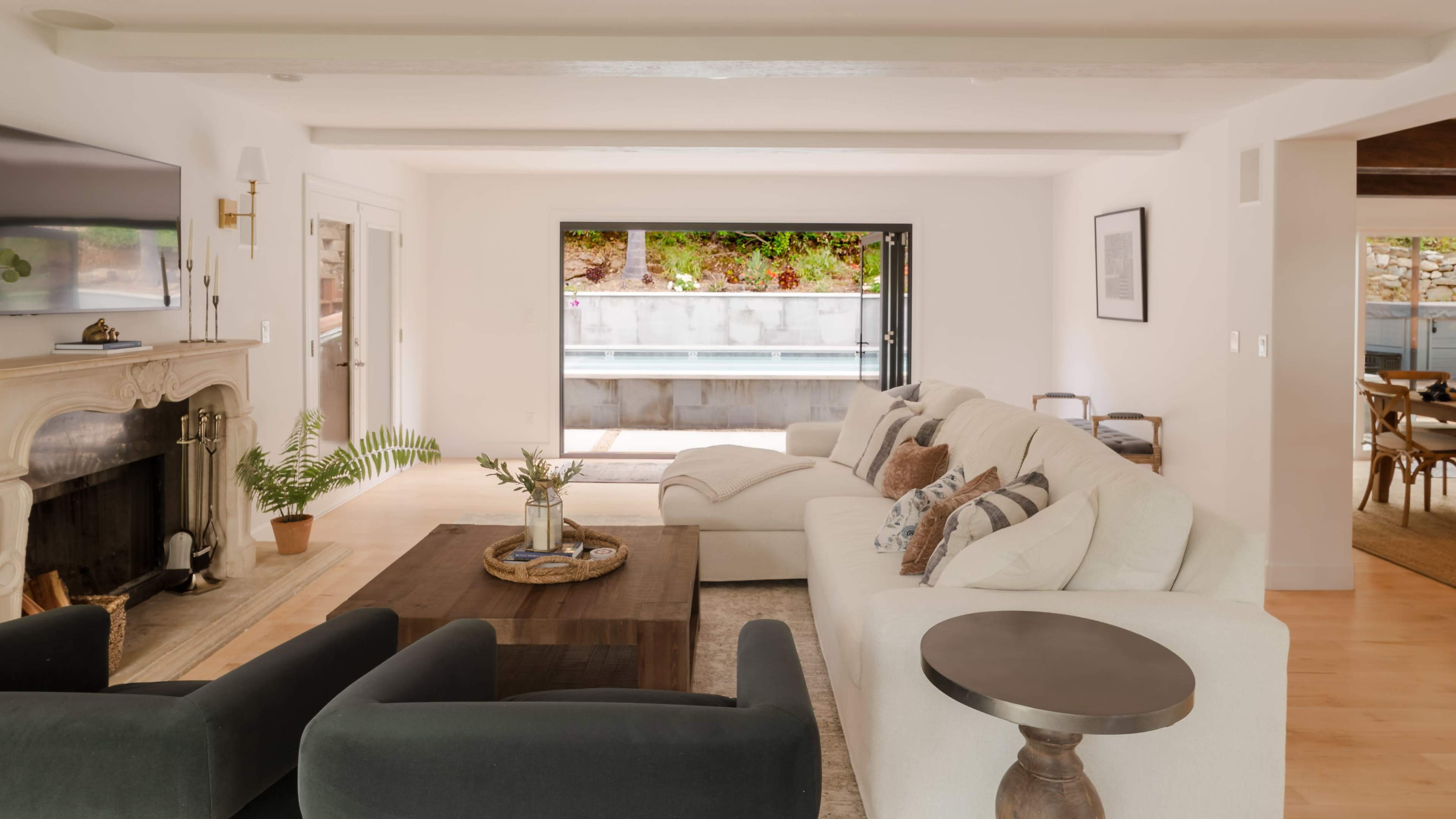 The image depicts a spacious, modern living room with a white sofa, dark chairs, and a coffee table, overlooking a pool area through large glass doors.