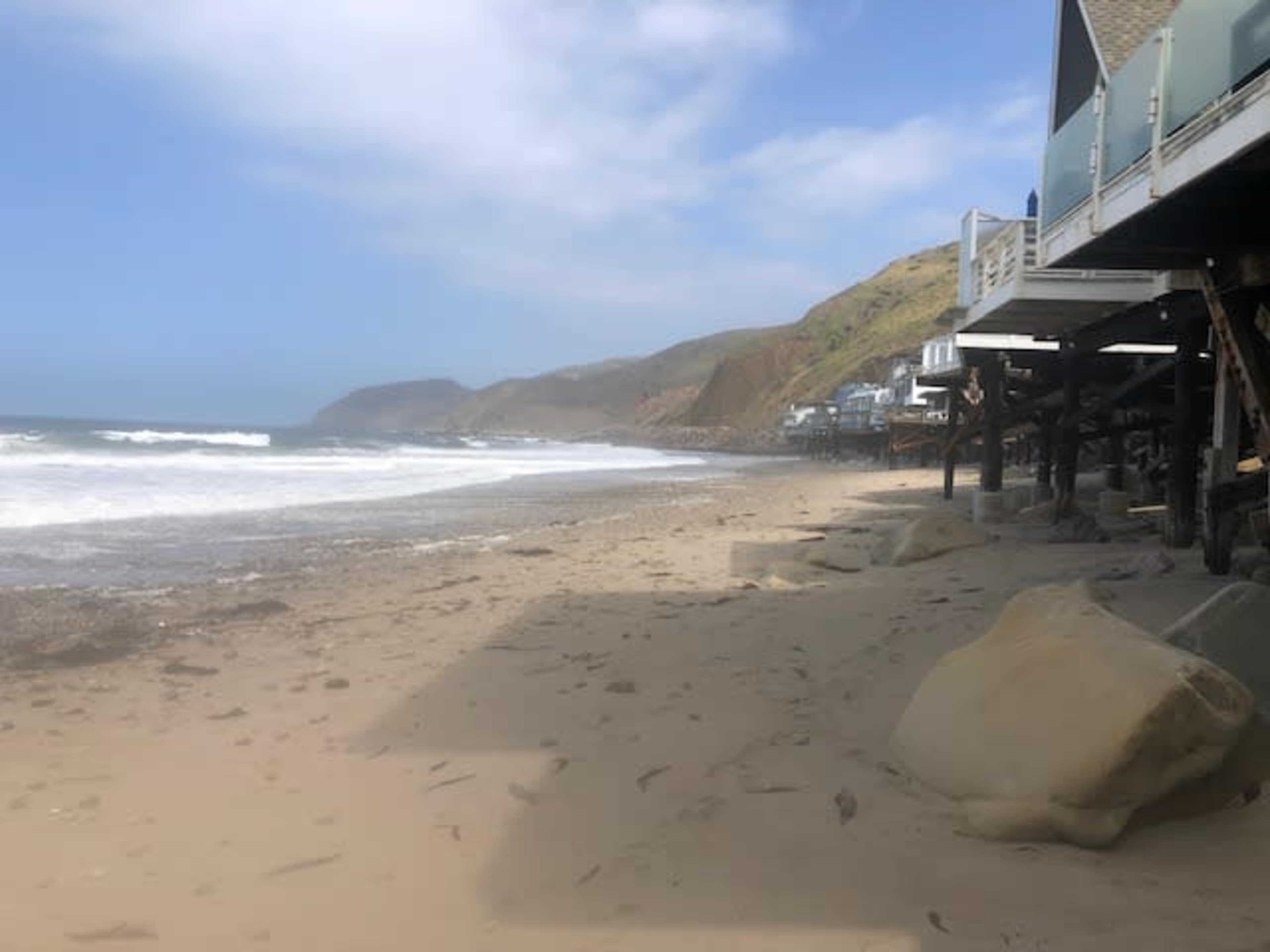 A sandy beach with waves lapping at the shore, flanked by rocky cliffs and structures built on stilts in the background.