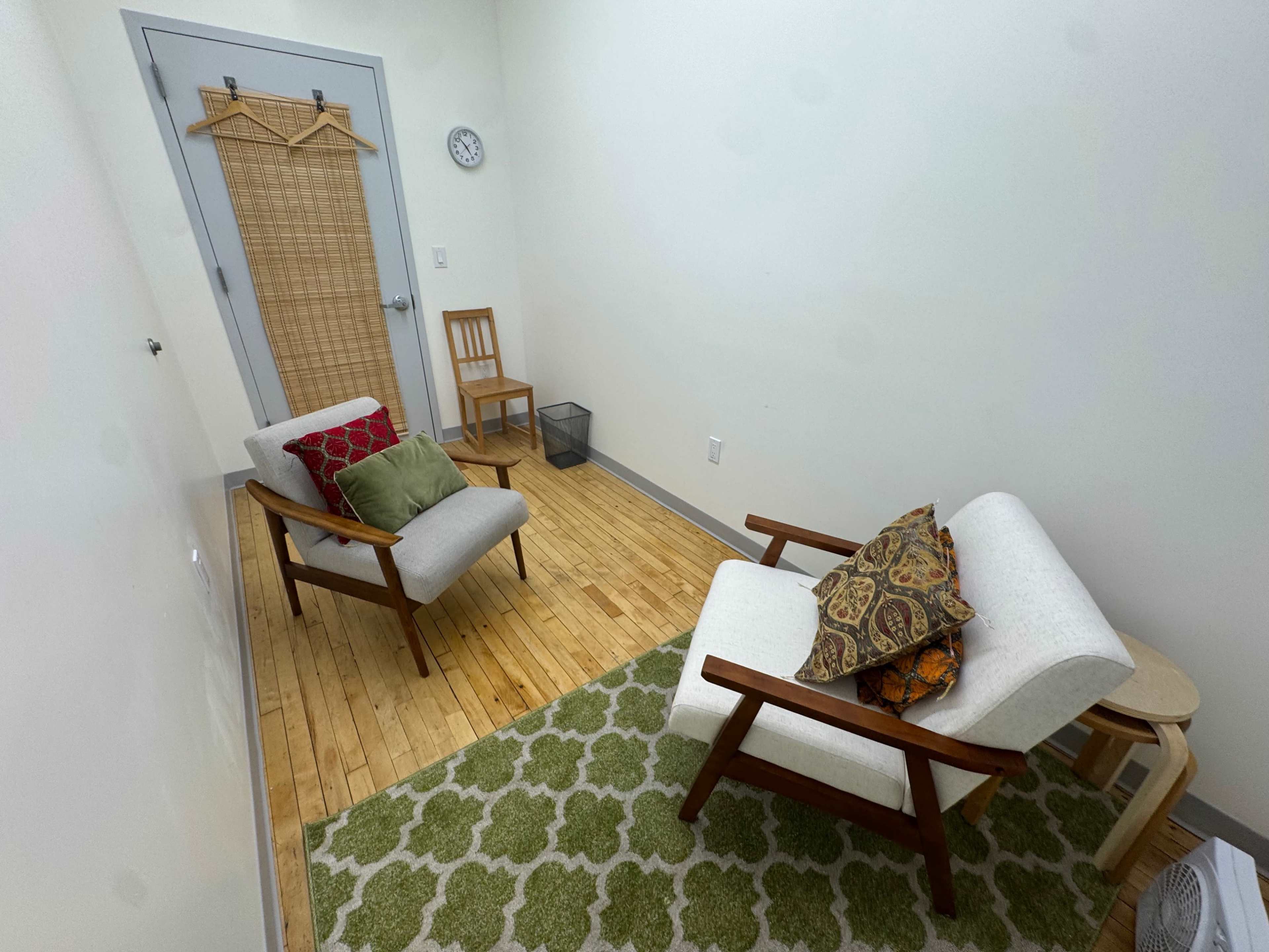 The image shows a small, minimalist room with two chairs, a small table, a rug, and a door.