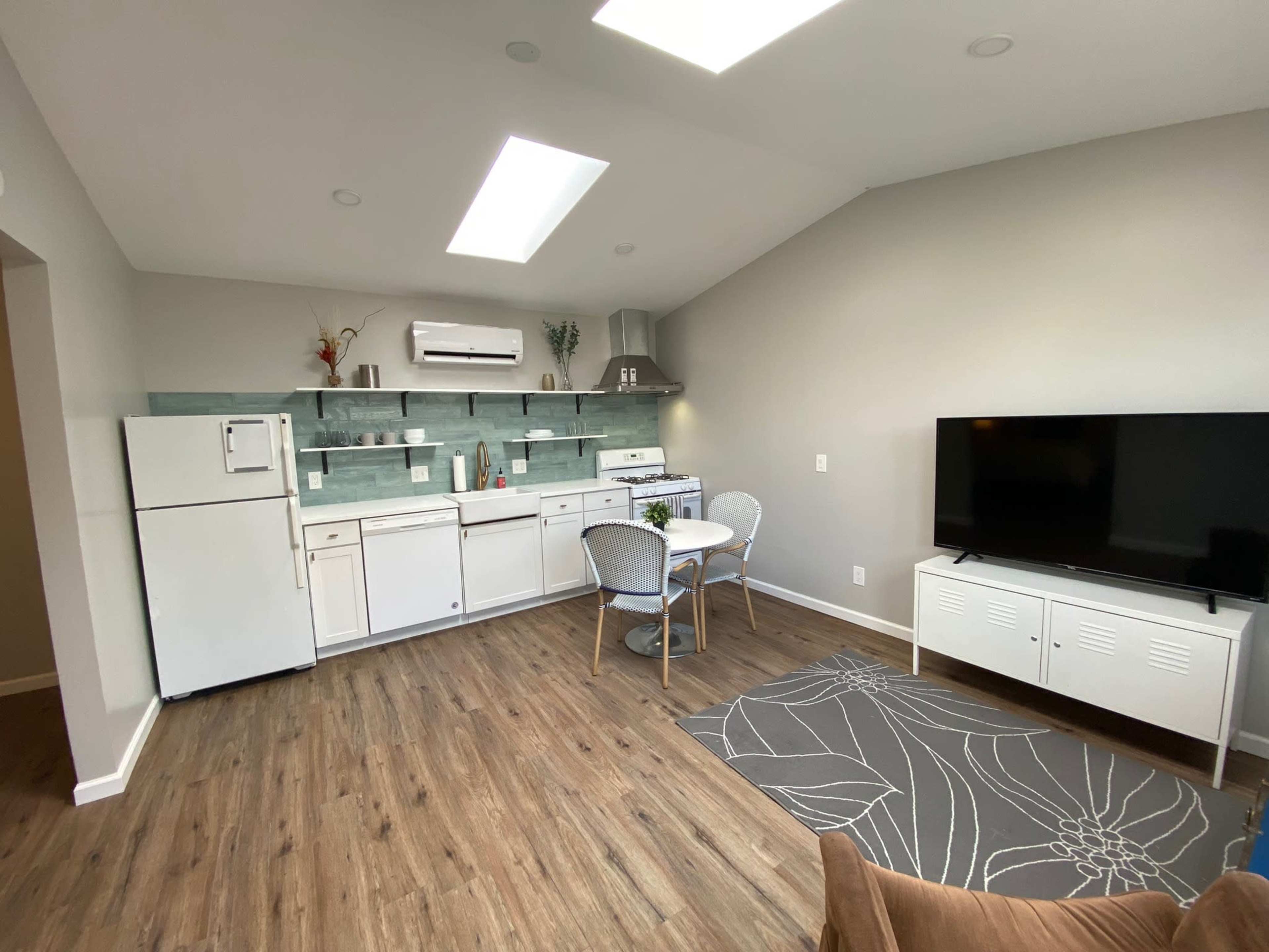 The image shows a compact kitchen and dining area featuring a refrigerator, stove, and a small table with two chairs, illuminated by skylights.
