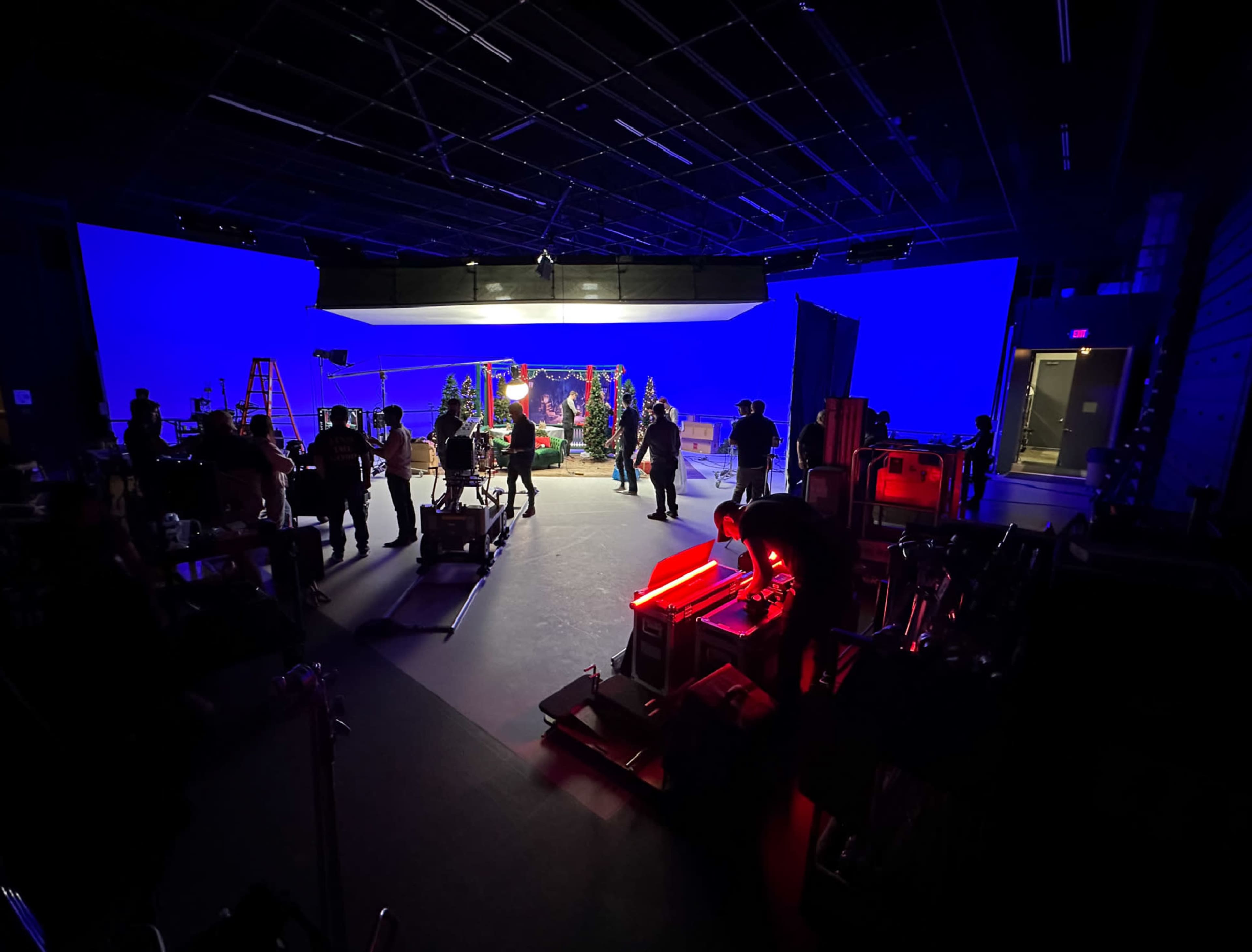 A film production crew sets up equipment in a studio, illuminated by blue background lighting and various tools scattered throughout the space.