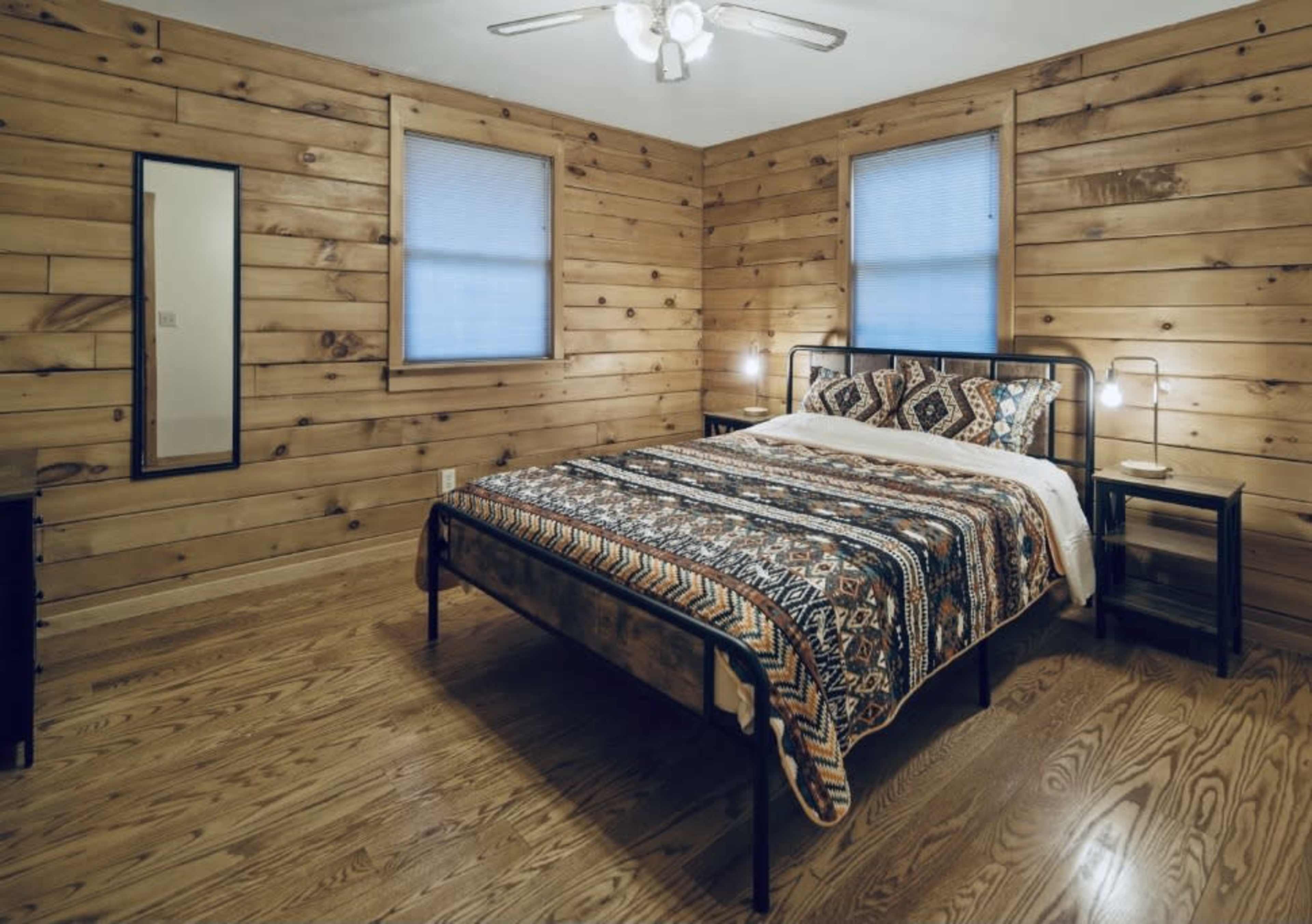 A simple bedroom features a metal bed frame with a patterned quilt, wooden walls, hardwood flooring, and two windows with blinds.