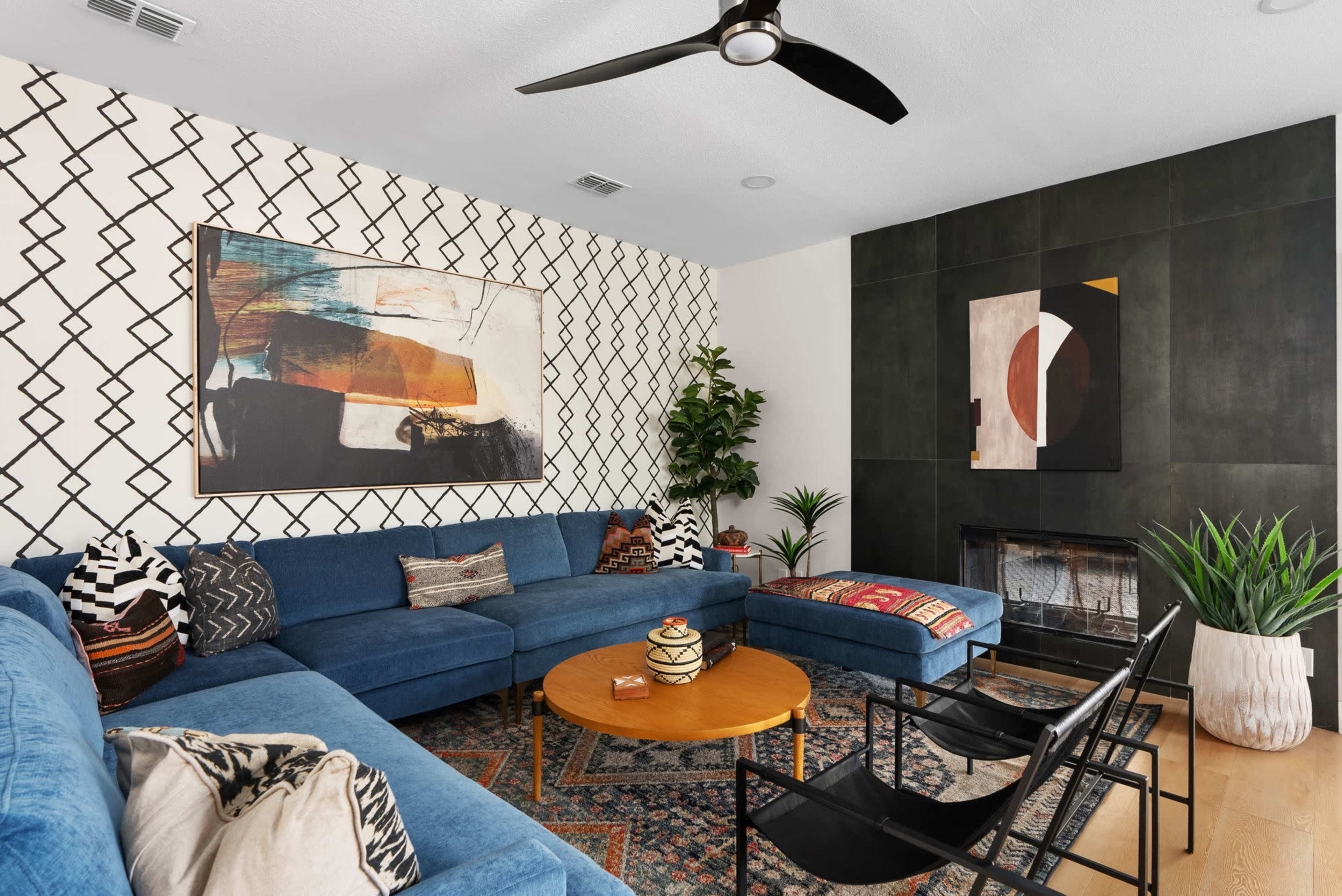 The image shows a modern living room featuring a blue sectional sofa, a round wooden coffee table, a patterned area rug, and a black fireplace, with abstract artwork on the walls.
