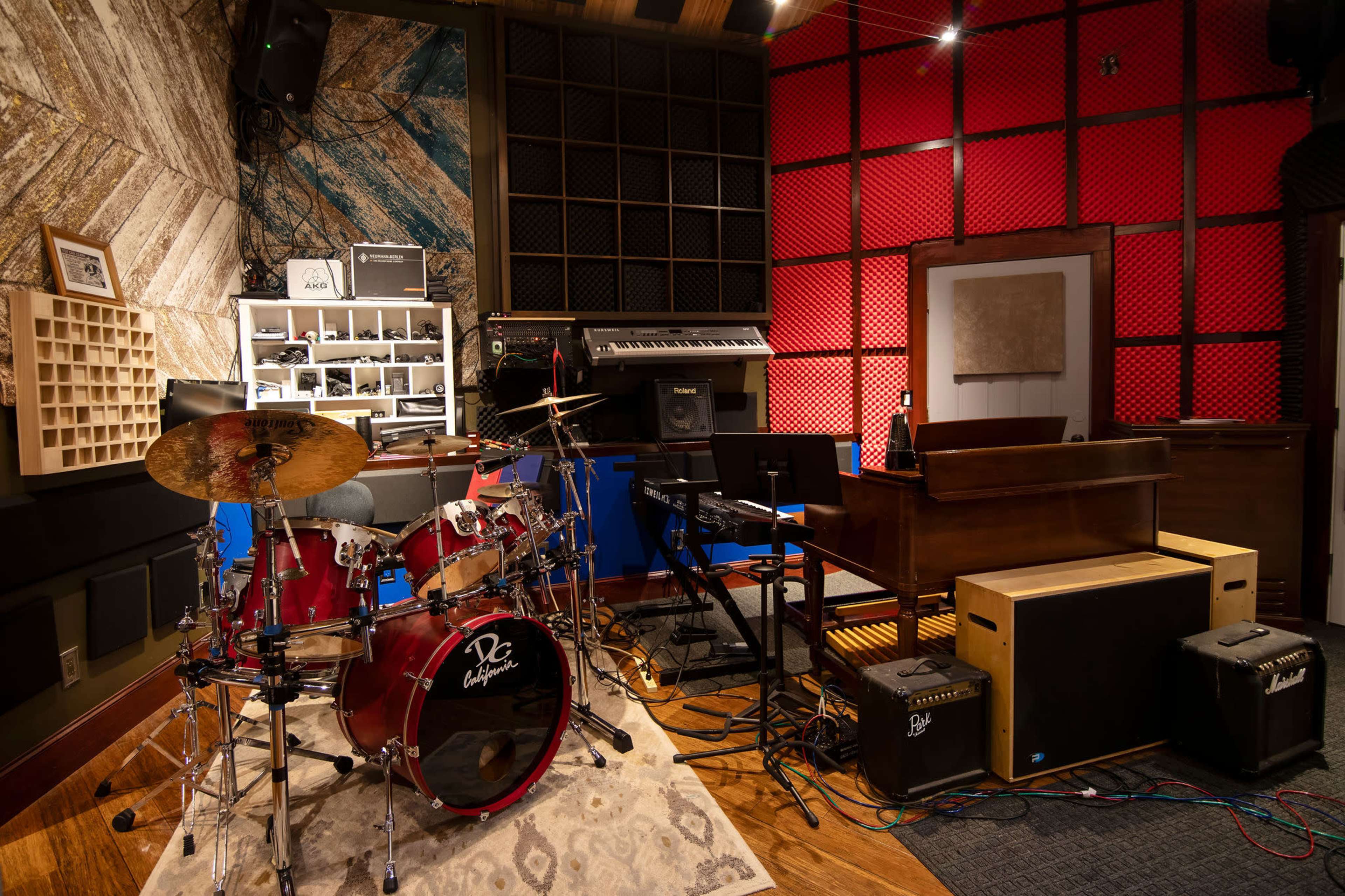 A music studio featuring a drum set, piano, keyboards, and various audio equipment arranged against a backdrop of textured walls.